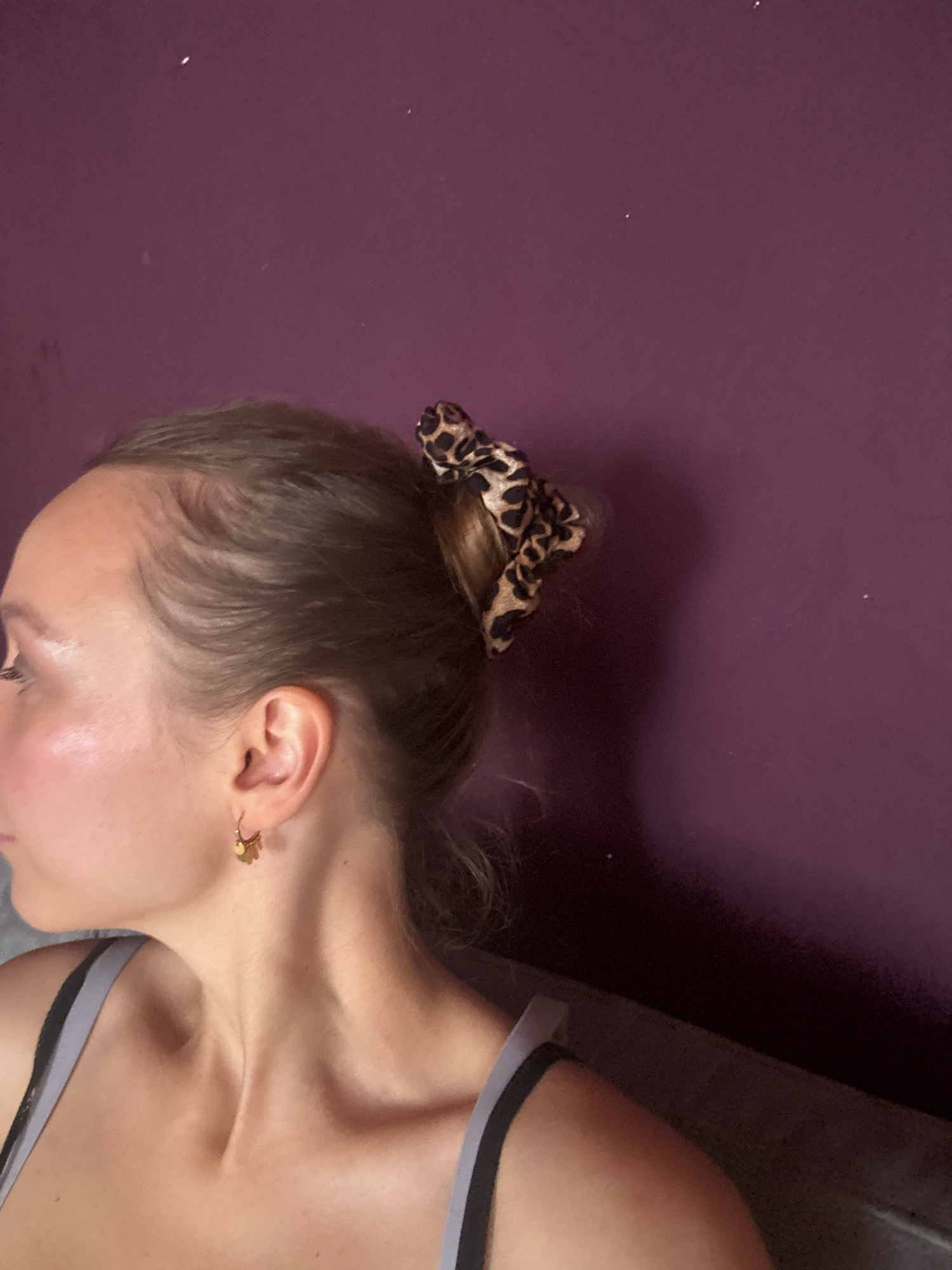 The image shows a close-up, profile view of the left side of a young woman's head and neck. She has light brown hair pulled up in a bun with a leopard print scrunchie. She is wearing a grey bra with black straps and a small gold earring in her left ear. The background is a solid purple wall with some small white specks. The lighting suggests an indoor environment. There is no discernible connection to Überherrn, Germany in the image.