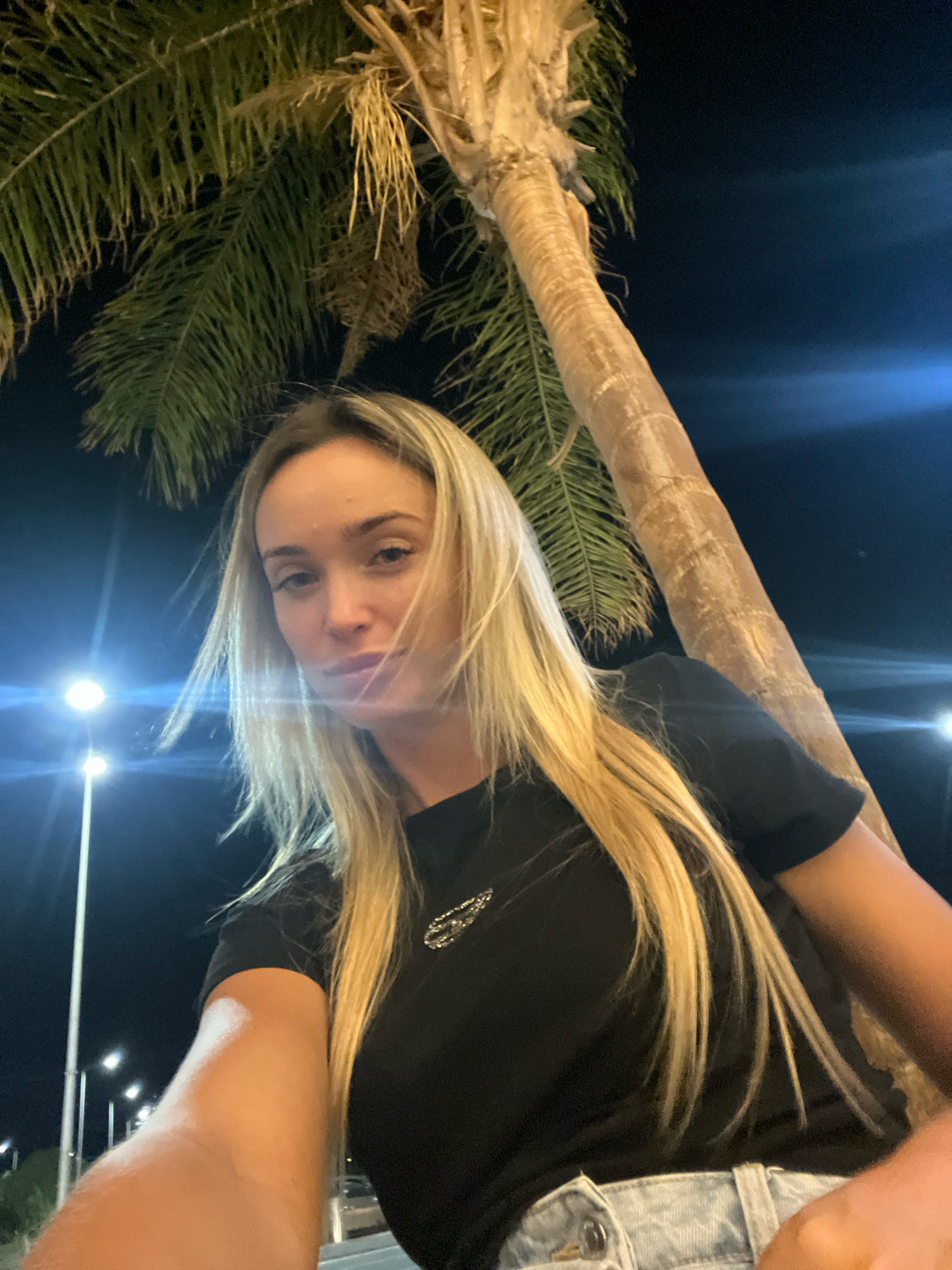 A young woman with long blonde hair is taking a selfie at night. She is wearing a black t-shirt and denim shorts. Behind her, a palm tree and streetlights are visible. The scene is likely in Sharm El Sheikh, Egypt, known for its beaches and nightlife.