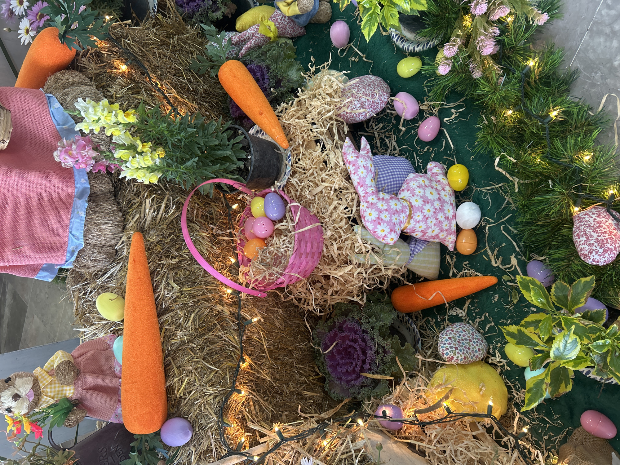 The image captures an elaborate Easter display, likely indoors. The scene incorporates elements of traditional Easter decor, including pastel-colored eggs, stuffed rabbits, and carrots, positioned among straw and artificial greenery. There are also plush scarecrows with straw hair and yellow dresses. Lights are intertwined within the display, creating a warm glow. There is no indication of its location in Puebla, Mexico.