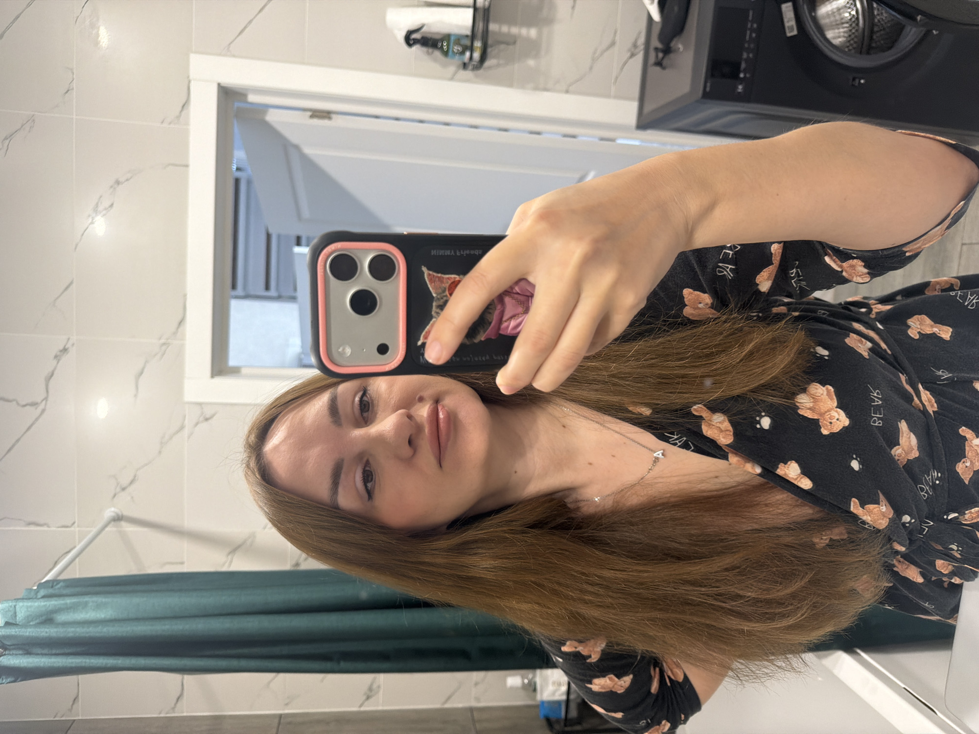 The image shows a woman taking a selfie in a bathroom. She is standing in front of a tiled wall and a dark green shower curtain. She has long, brown hair and is wearing a black nightgown with a teddy bear print. She is holding her phone in her right hand, and her left hand is on her hip. In the background, there is a washing machine and a shelf with toiletries. The overall setting appears to be a domestic bathroom.