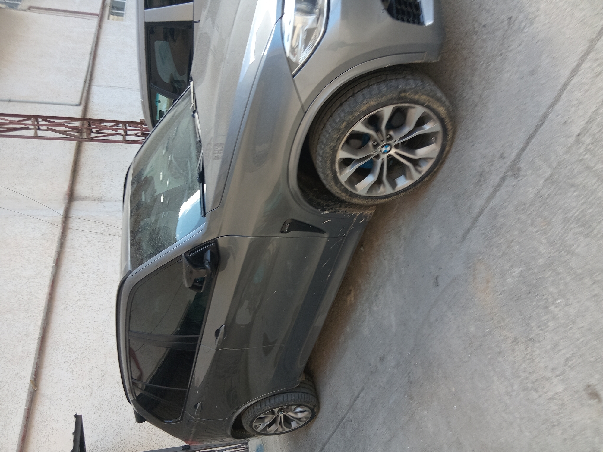 The image shows a gray BMW X5 SUV parked on a gray concrete surface. The vehicle is angled in the frame. The front passenger-side wheel is visible in detail, with a multi-spoke design and a BMW logo. The front headlight is also visible. The body of the SUV has some dirt and streaks.
In the background, a light-colored wall is visible, with metal support structures attached to it. Windows can be seen in the wall. The setting appears to be an outdoor parking area. The city of Batumi, Georgia is not evident from the image.