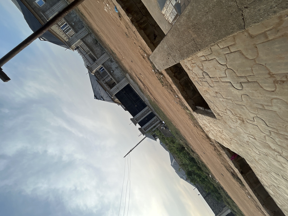 An urban landscape under a cloudy sky features buildings in varying stages of construction. One building close up has a stone-like facade with a repeating decorative pattern. A dirt road or path runs parallel, with additional buildings visible beyond. A pole extends upwards into the sky, adding a vertical element to the scene. The entire view is rotated clockwise.