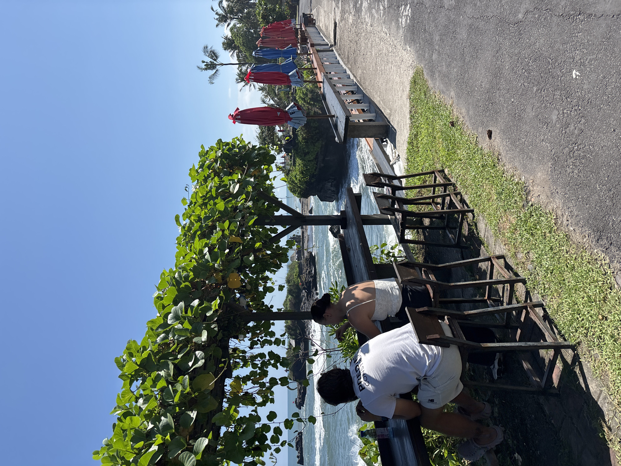 A sunny day in Mengwi, Indonesia, features a coastal scene. Two people lean against a weathered, dark-colored metal railing overlooking a rocky coastline where waves crash. The person on the left wears a white tank top, while the person on the right sports a white t-shirt with visible text and light-colored shorts. Behind them, a paved path runs adjacent to a grassy area. A row of colorful umbrellas, predominantly red and blue, lines a slightly elevated platform. The sky is clear and blue. Lush green foliage frames the view, adding a tropical feel to the location.