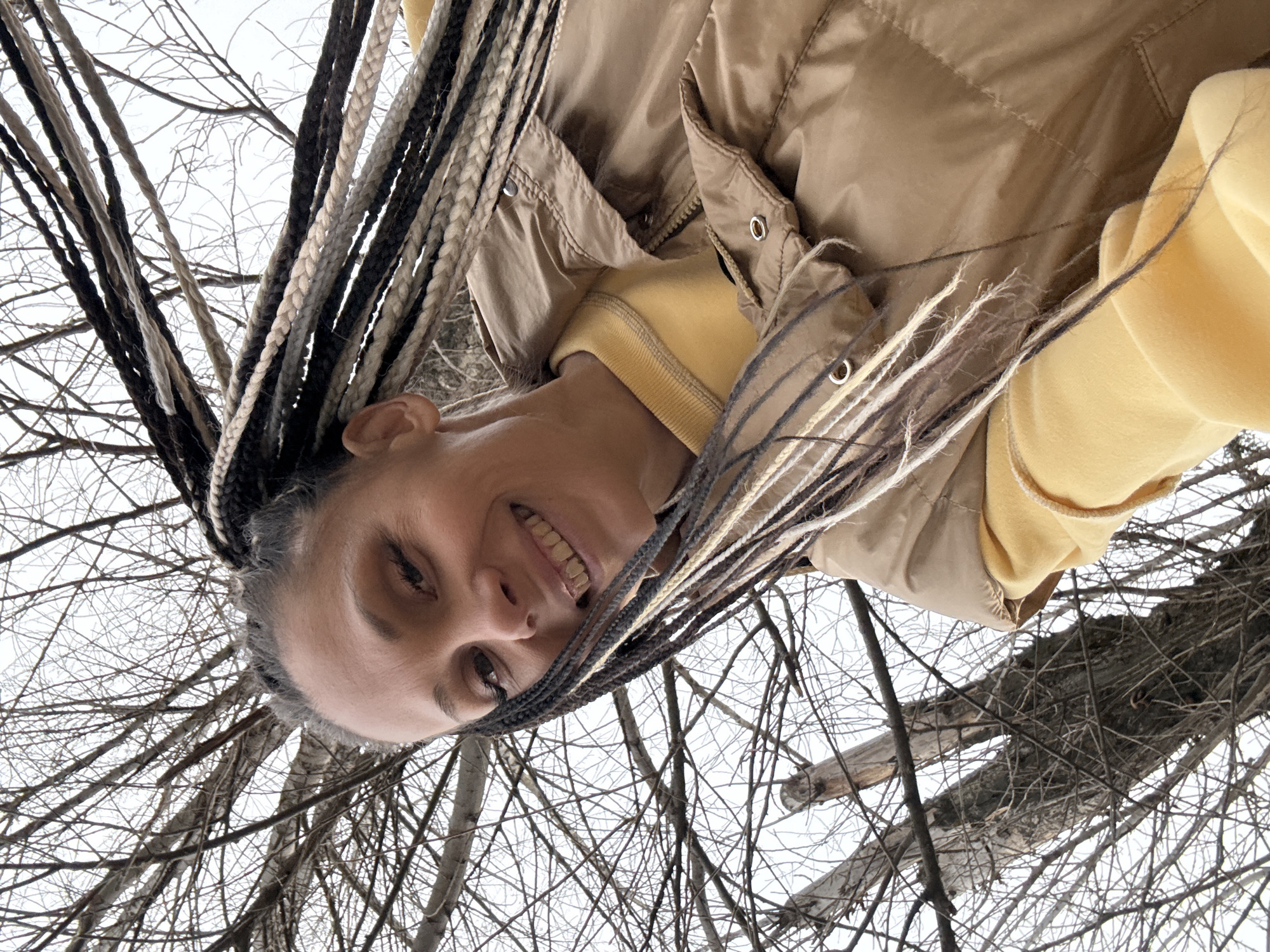 A woman with braided hair smiles at the camera, which is positioned from a low angle looking up at her. Her hair is a mix of black, grey, and white braids. She is wearing a light brown puffy vest over a yellow sweater. Behind her and slightly out of focus are bare tree branches against a white sky. The location is presumably a wooded area in Rusanovo, Russia.