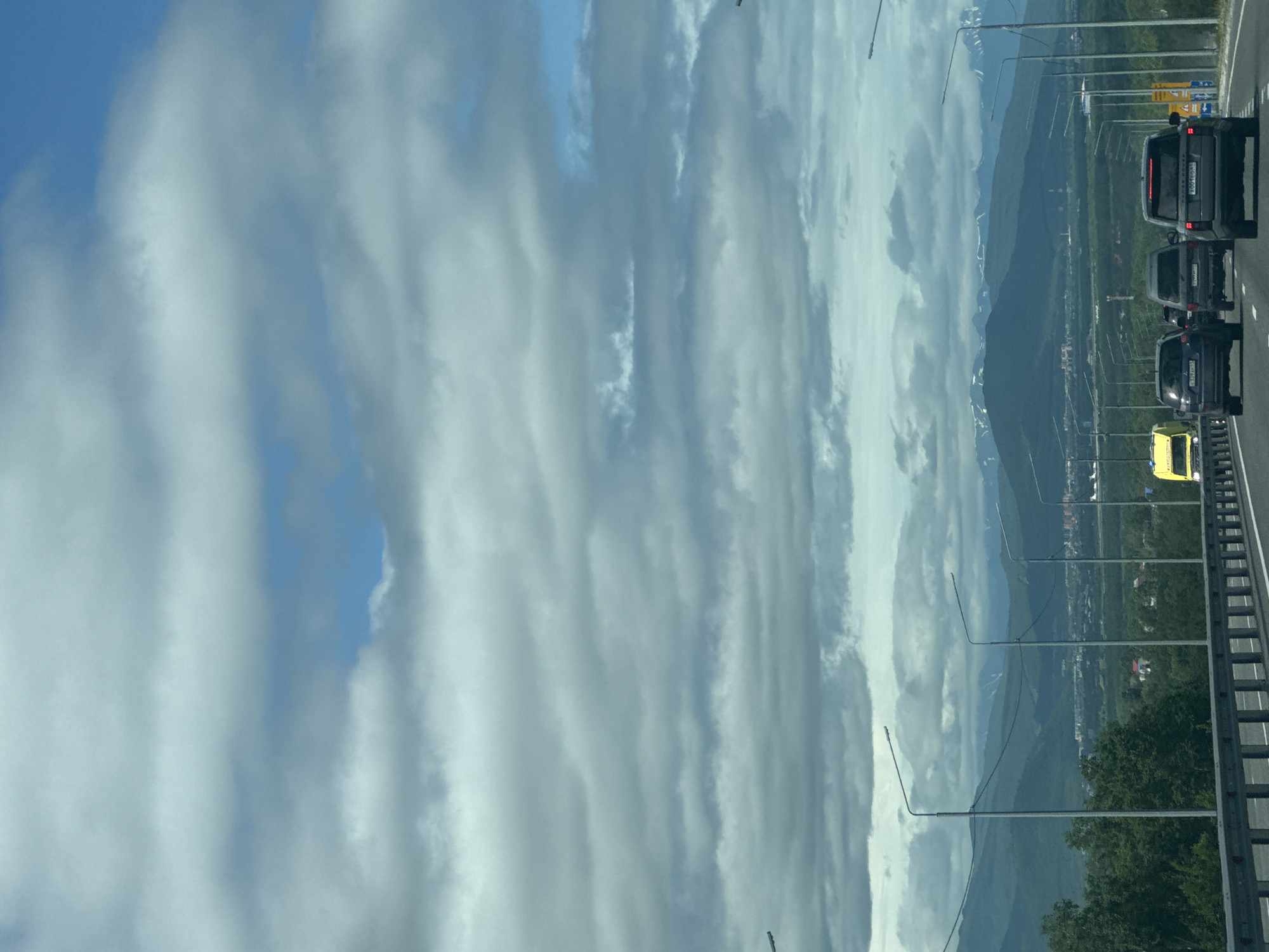 The image depicts a highway scene with a mountainous landscape in the background. Several vehicles are visible on the road, including SUVs and a yellow truck. The sky is filled with a mix of clouds and patches of blue. Streetlights line the side of the highway. In the distance, a city or town is nestled at the foot of green, rolling hills, with snow-capped mountains rising behind them. The weather appears overcast but with potential for sunlight breaking through.