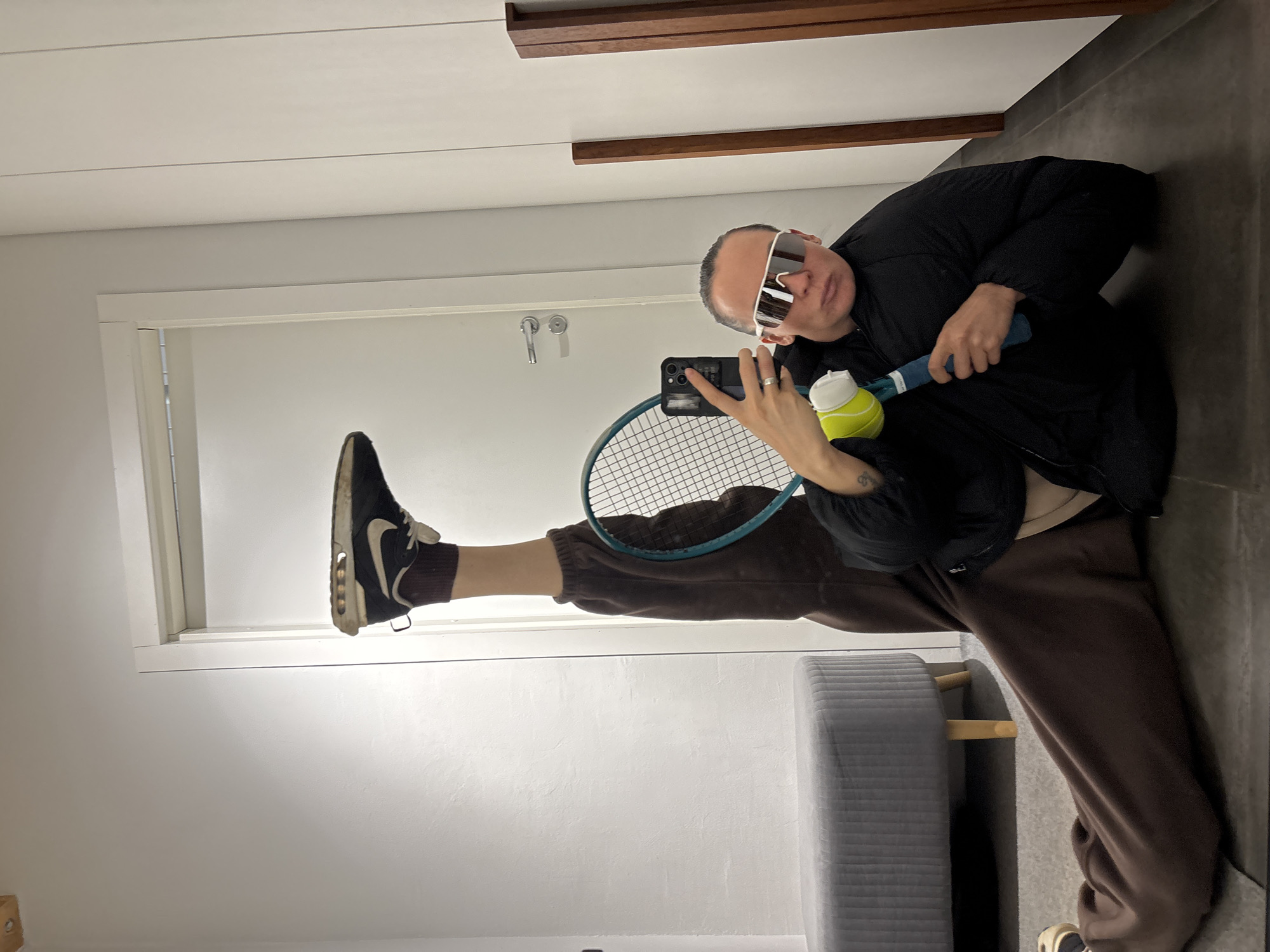 A person wearing sunglasses, a black jacket, and brown sweatpants holds a tennis racket and tennis ball while lying on their side. Their left leg is raised, with a black and white sneaker touching the door frame. They are holding a cell phone in their right hand, apparently taking a selfie. The background includes a white door, light gray walls, and a gray padded bench with wooden legs. The ceiling is white with a brown decorative strip. The floor is dark gray tile. This scene could take place in a modern interior with a focus on athletic activity. The location in the city of Chaikovsky, Russia is undetermined.