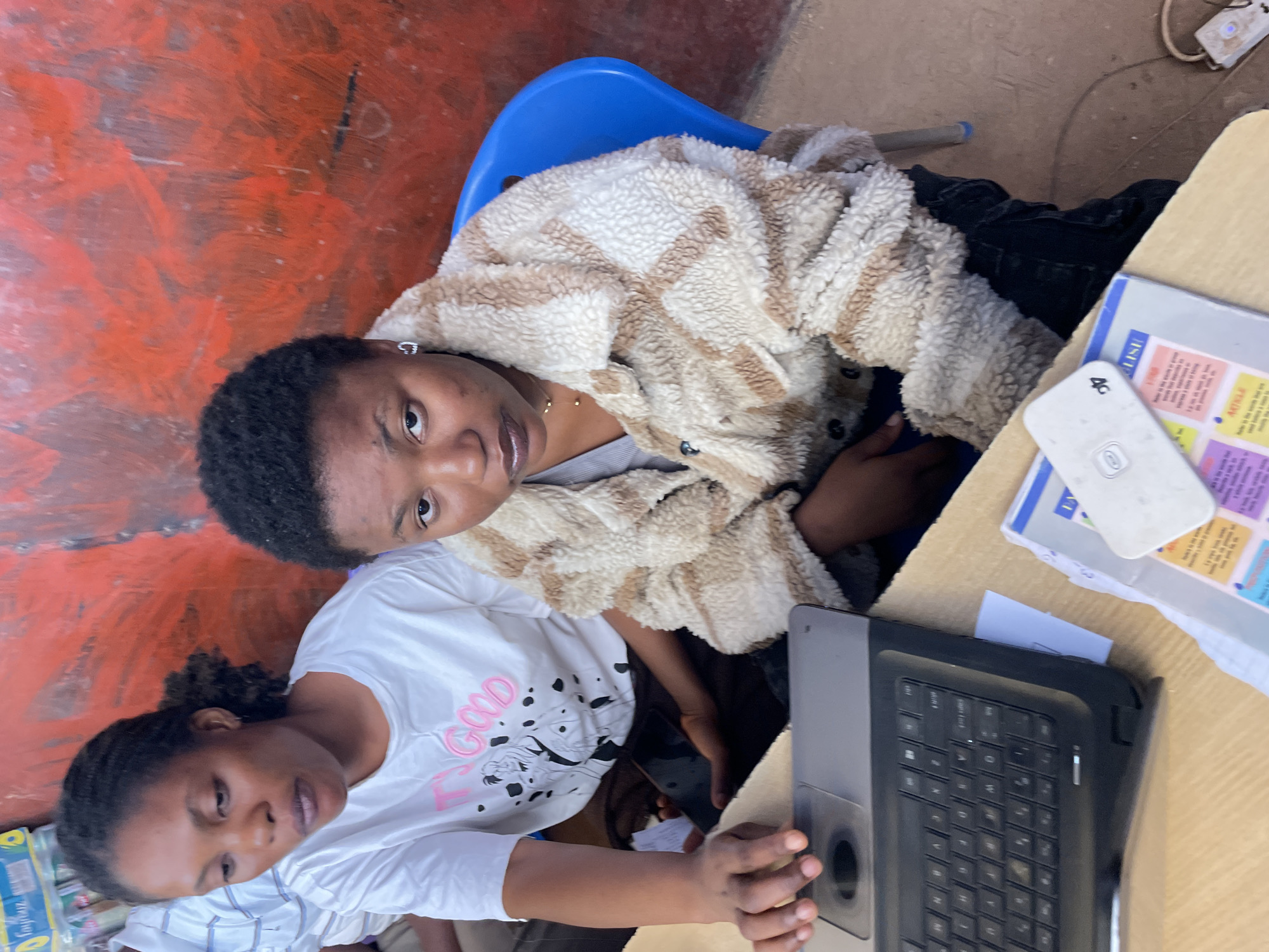 Two young women are sitting at a desk, working on a laptop. One woman is wearing a white shirt with a pink "good" inscription. The other woman is wearing a white and brown jacket. A textbook and a white device are on the desk. The location is Gura Top, Nigeria. The wall is painted orange.