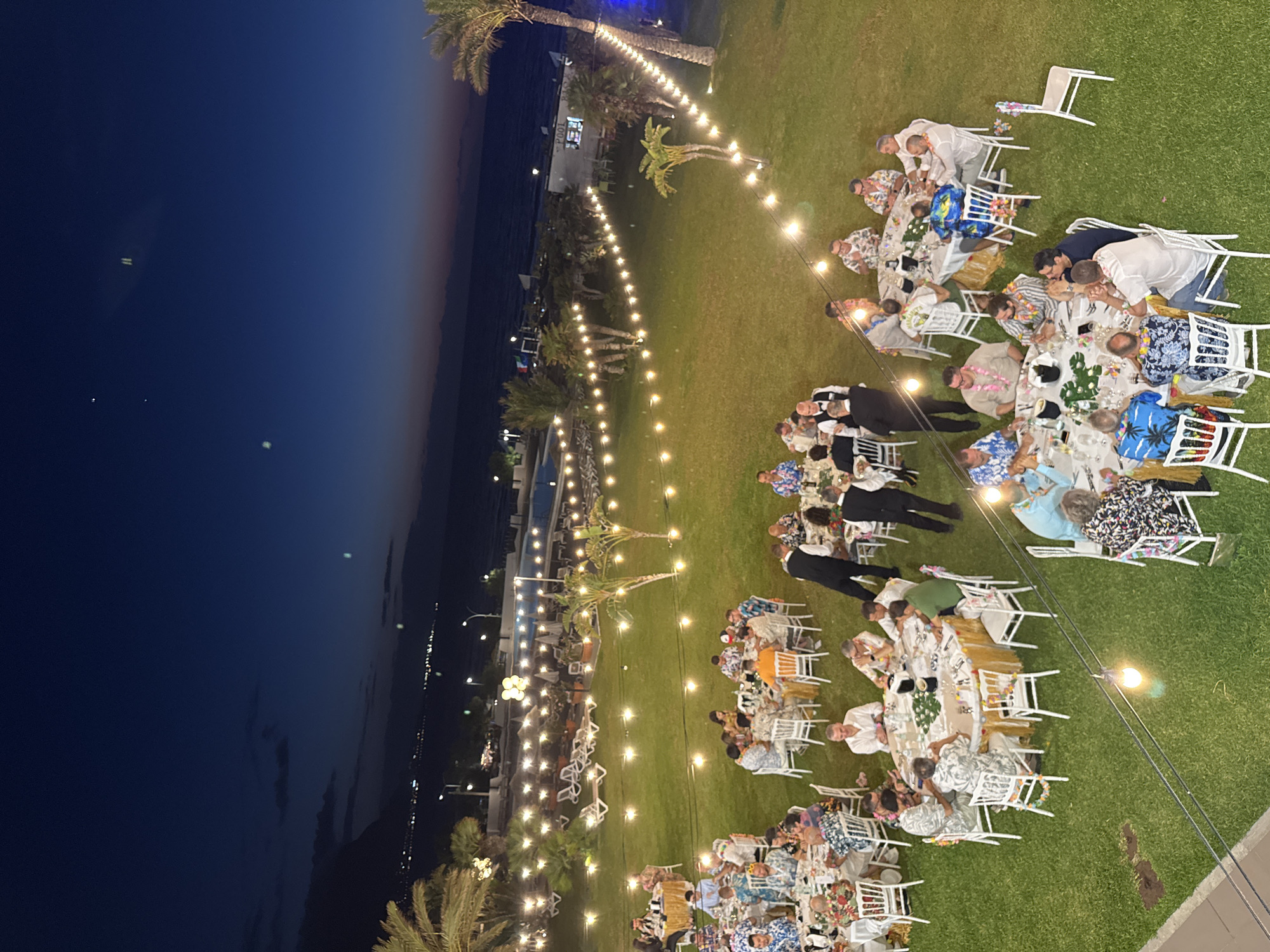 An outdoor evening event is taking place on a grassy area adjacent to a body of water, possibly the sea, with lights from a distant city visible on the horizon. The scene is illuminated by string lights and table lamps. Several round tables are set up for a gathering, with white chairs arranged around them. Many of the attendees are wearing Hawaiian-style shirts. Some tables are decorated with flowers and greenery. In the background, there is a swimming pool and lounge chairs, suggesting a resort or hotel setting. The sky is a deep blue, transitioning to a lighter hue near the horizon, indicating dusk or twilight. The overall atmosphere appears festive and social. The location is the city of Isola delle Femmine, Italy.