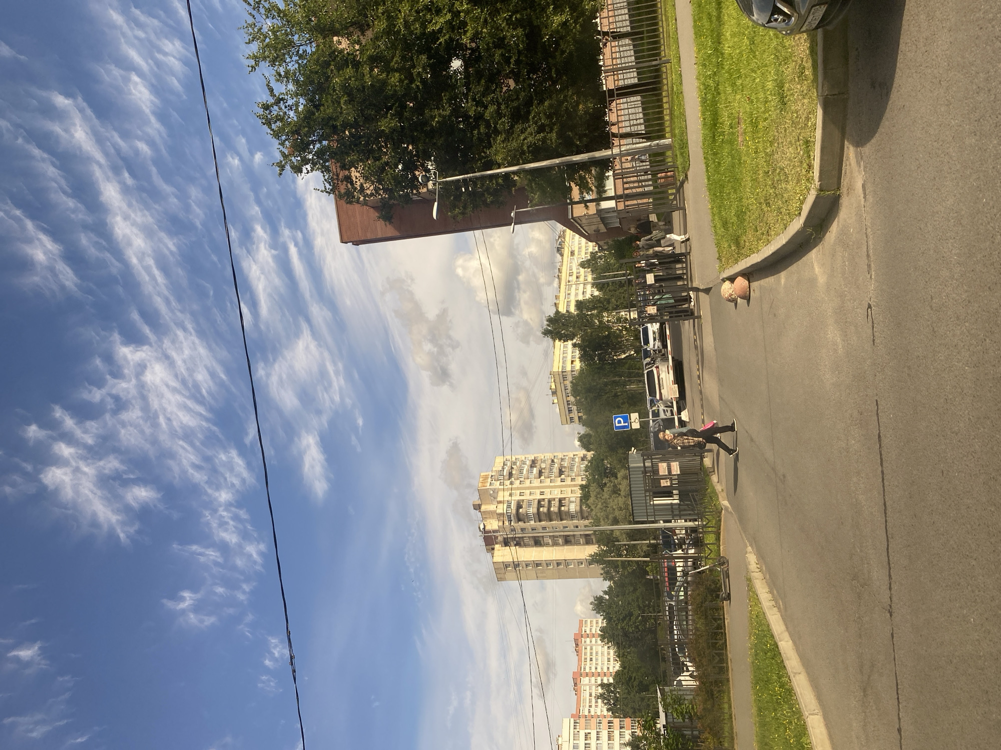The image depicts a street scene in Saint Petersburg, Russia, on a day with a clear blue sky featuring wispy white clouds and some scattered larger clouds. In the foreground, an asphalt road curves to the right, bordered by a grassy verge with a concrete curb. A woman wearing a plaid shirt and dark pants is crossing the road from left to right, carrying a pink bag. To her left, an electric scooter is parked on the grass.
Beyond the road, a black metal fence encloses a parking area where several cars are visible, including a white SUV. A blue parking sign with a white "P" is mounted on a post near the entrance to the parking lot, adjacent to a pedestrian crossing. Several people are standing near the entrance gate.
Further in the background, rows of multi-story residential buildings are visible. The tallest building in the mid-ground is a light-colored, mid-rise apartment building with numerous balconies. To its left, a lighter-colored apartment building with orange and white sections is visible, and to its right, another apartment building with light-colored facades and a yellow section. Lush green trees are scattered throughout the scene, with a large, dense tree dominating the right side of the image, partially obscuring a tall, reddish-brown brick building. Overhead, power lines are strung across the sky. The overall impression is a typical urban residential area.