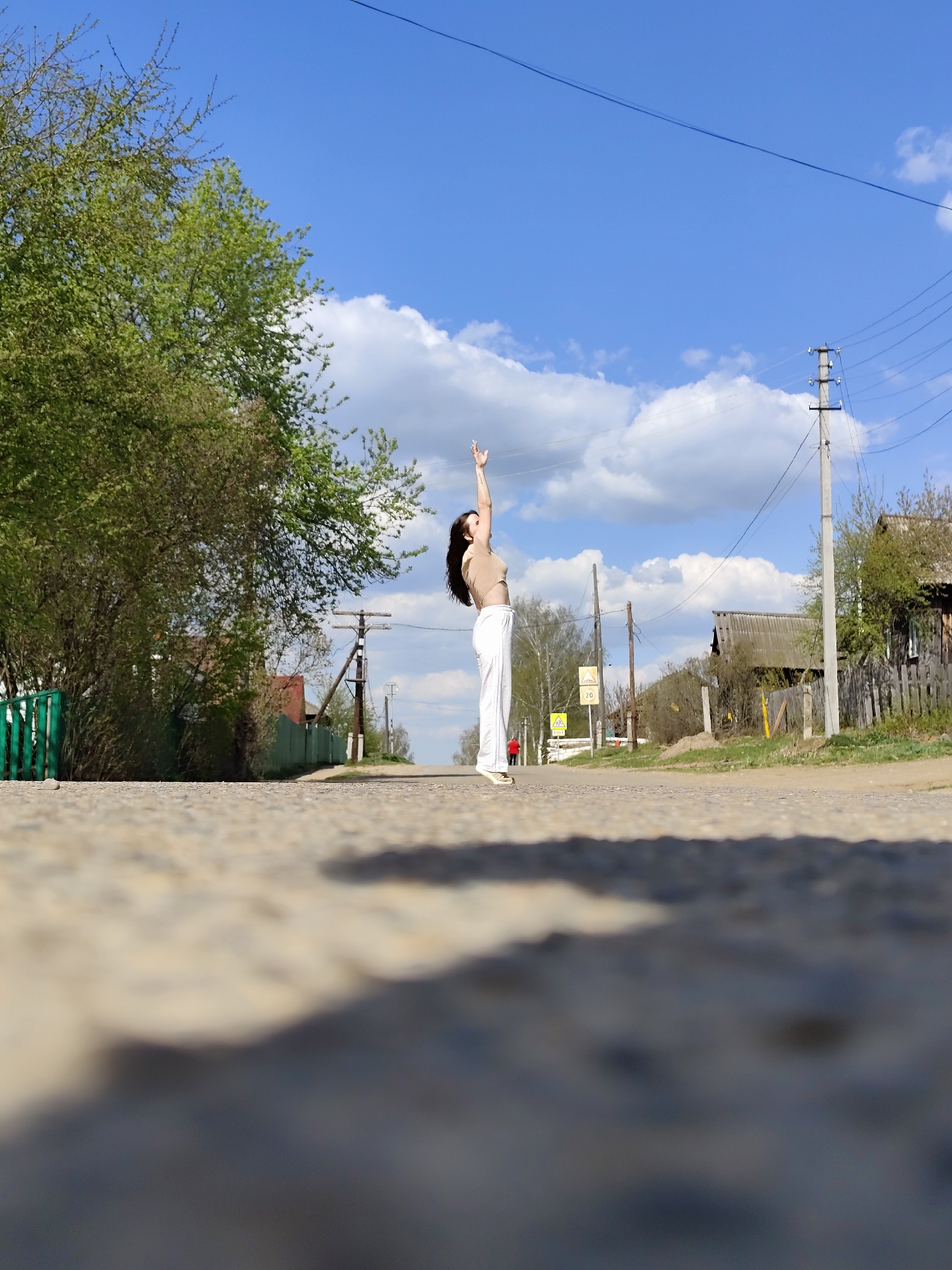 A woman stands on an asphalt road with her arms raised, likely in Фоки, Russia. She is wearing a beige top and white pants, and her dark hair is long. The road stretches away from the viewer, flanked by greenery on the left and wooden structures and utility poles on the right. The sky is blue with scattered white clouds. The perspective is from a low angle, blurring the foreground of the road.
