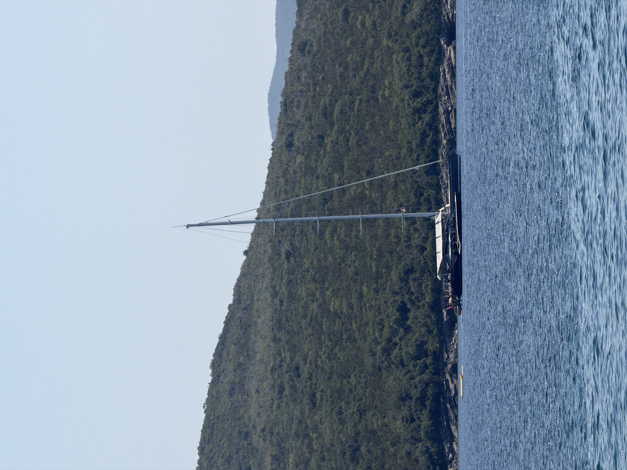 The image shows a large sailboat anchored in the bay of Bigova, Montenegro. The sailboat has a tall mast and a white sail. The bay is surrounded by a lush green hillside. The water is a deep blue and appears calm. There is a small yellow raft floating near the sailboat. The sky is clear and blue.