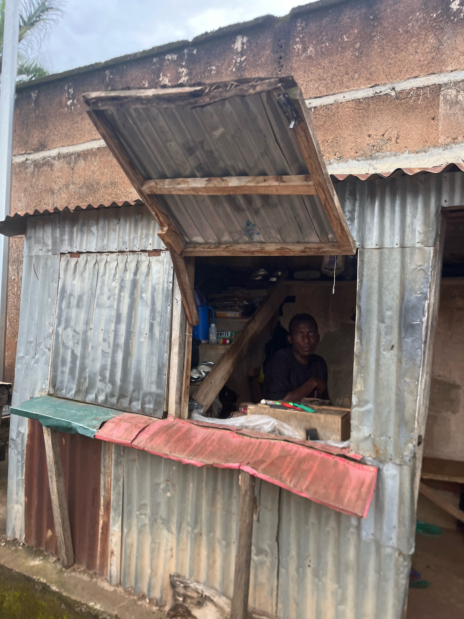 The image shows a man sitting inside a small, makeshift shop in the city of Jos, Nigeria. The shop appears to be constructed from corrugated metal sheets and has a wooden counter. The man is wearing a dark t-shirt and is looking towards the camera. There are some items on the counter in front of him, including pens and what appear to be some packaged goods. Behind him, there are shelves with various items, including bottles and papers. The overall environment appears somewhat unkempt, with signs of wear and tear on the structure. The sky in the background suggests an overcast day.
