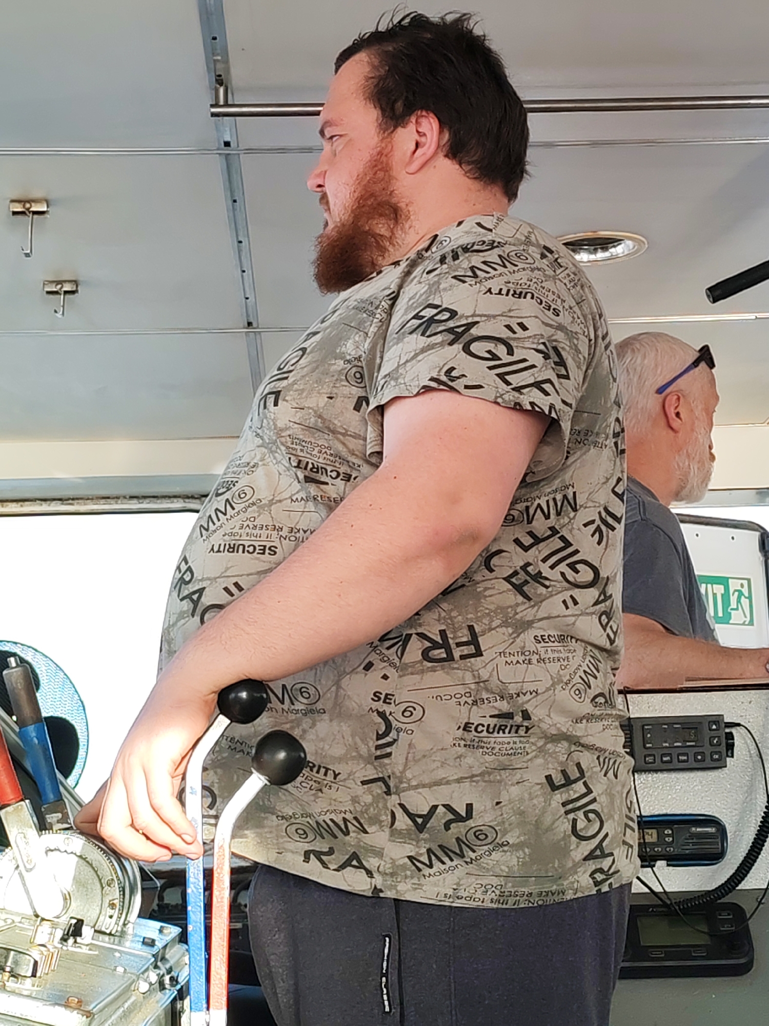 The image shows two men inside what appears to be a control room of some kind. The primary subject is a man with a reddish beard, wearing a patterned t-shirt with words like "FRAGILE" and "SECURITY" printed on it. He is operating levers on a piece of machinery. Behind him, a second man with white hair and sunglasses on his head is visible near electronic equipment. The interior has a utilitarian appearance, with metallic elements and what appear to be communication devices. The location of the scene is unknown.