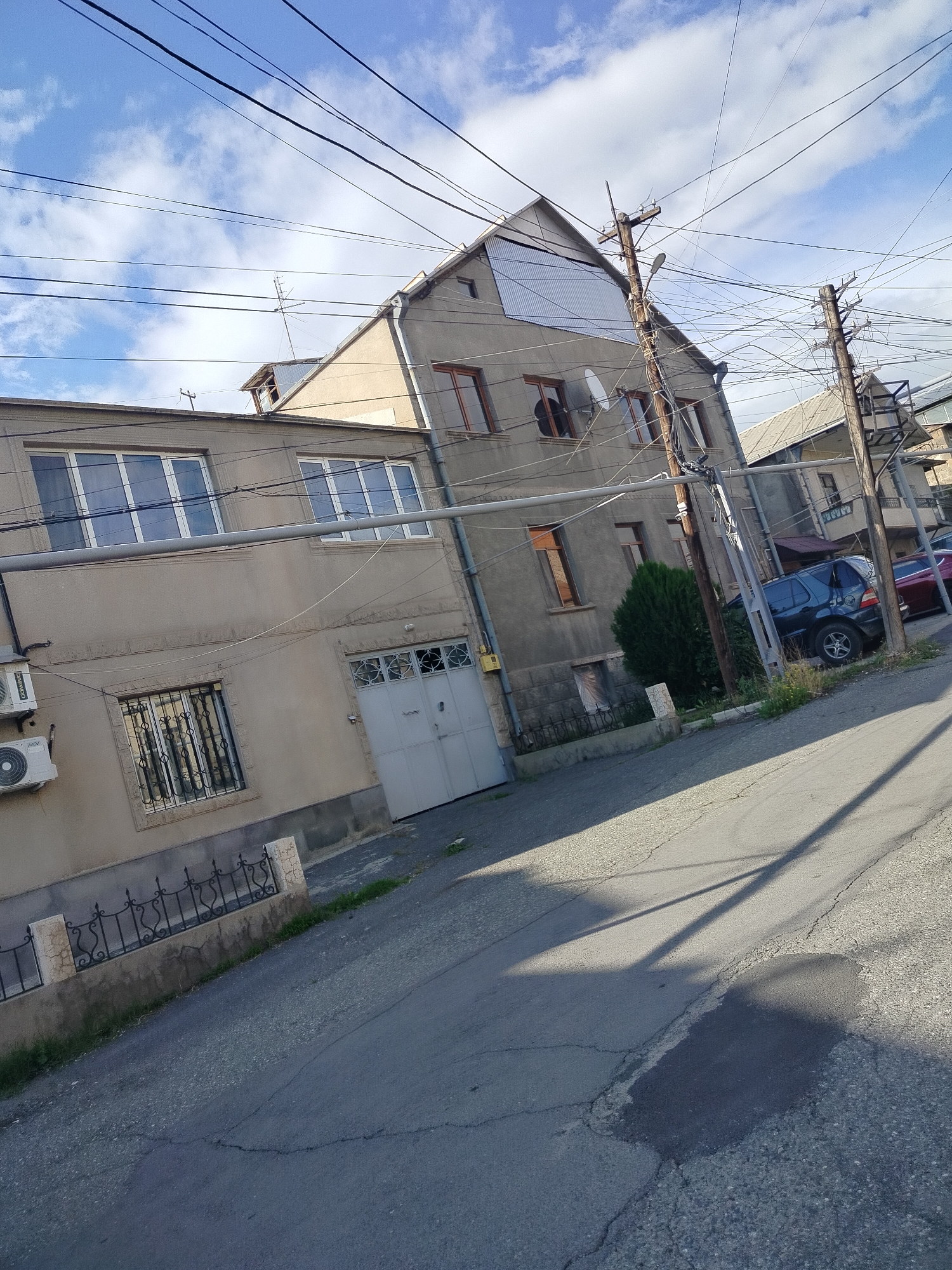 The image depicts a street scene in Yerevan, Armenia. The foreground consists of a cracked asphalt road with a shadow cast across it. To the left, there is a two-story residential building with light-colored stucco walls. This building features several windows, some with decorative metal bars, and a closed metal garage door. An air conditioning unit is mounted on the exterior wall of the first floor. A short, decorative iron fence runs along the edge of the property, separating it from the sidewalk.
To the right of the two-story building is a taller building, possibly an older structure, with a grey facade. Its windows are rectangular. Power lines crisscross above the street. Three utility poles are visible, aligned along the street.
Cars are parked along the edge of the street, adjacent to the buildings. A small evergreen shrub is located near the base of one of the buildings. The sky is blue with scattered white clouds.