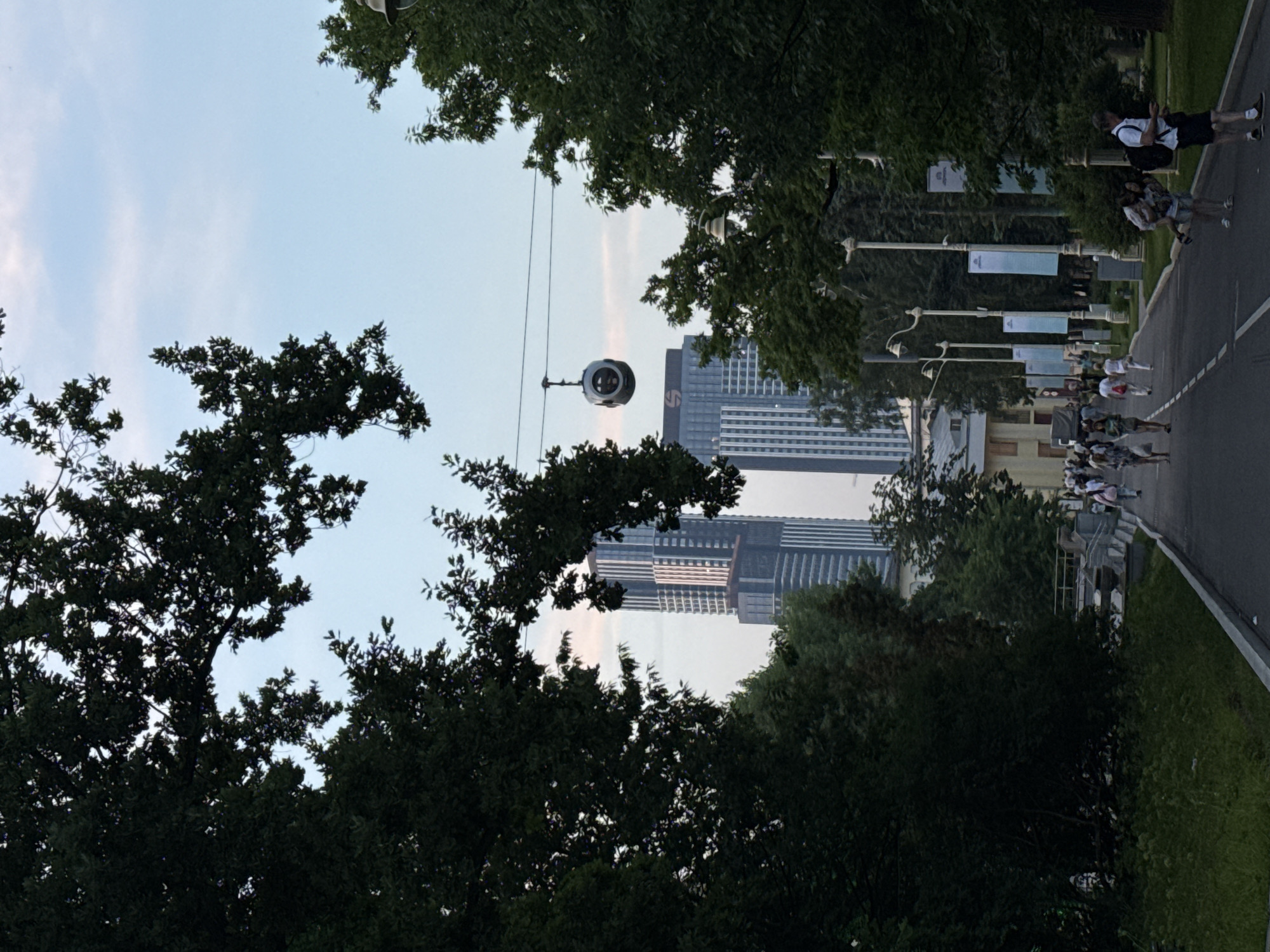 The image shows a tree-lined path in a park-like setting, with tall buildings in the background. A cable car is suspended overhead. Several people are walking along the path, some in small groups, others individually. The path is paved and bordered by grass and trees. In the distance, modern high-rise buildings, typical of Moscow's skyline, are visible. The sky is clear with some light clouds. This scene appears to be within the VDNKh (Exhibition of Achievements of National Economy) park in Moscow, Russia, which is known for its grand pavilions, fountains, and green spaces, as well as modern architectural elements interspersed with historical ones.