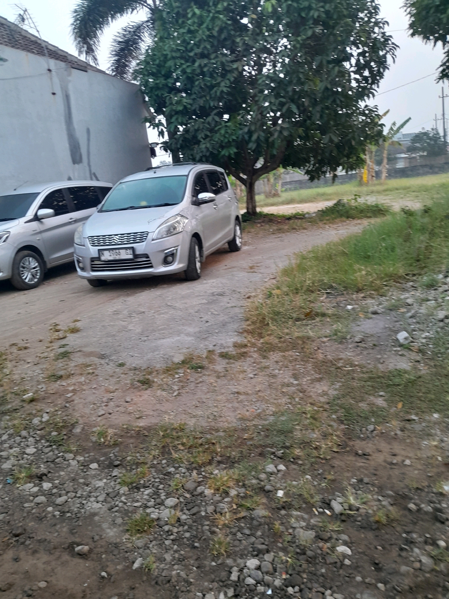 Two silver cars are parked on a dirt road in Glanggang, Indonesia. A silver sedan is parked on the left, and a silver minivan is parked on the right. The minivan has a license plate that reads "N 1366 GJ". A large tree with green leaves is behind the minivan, and a white building is behind the sedan. The ground is covered with dirt and gravel. There are some weeds growing on the side of the road. The sky is overcast.