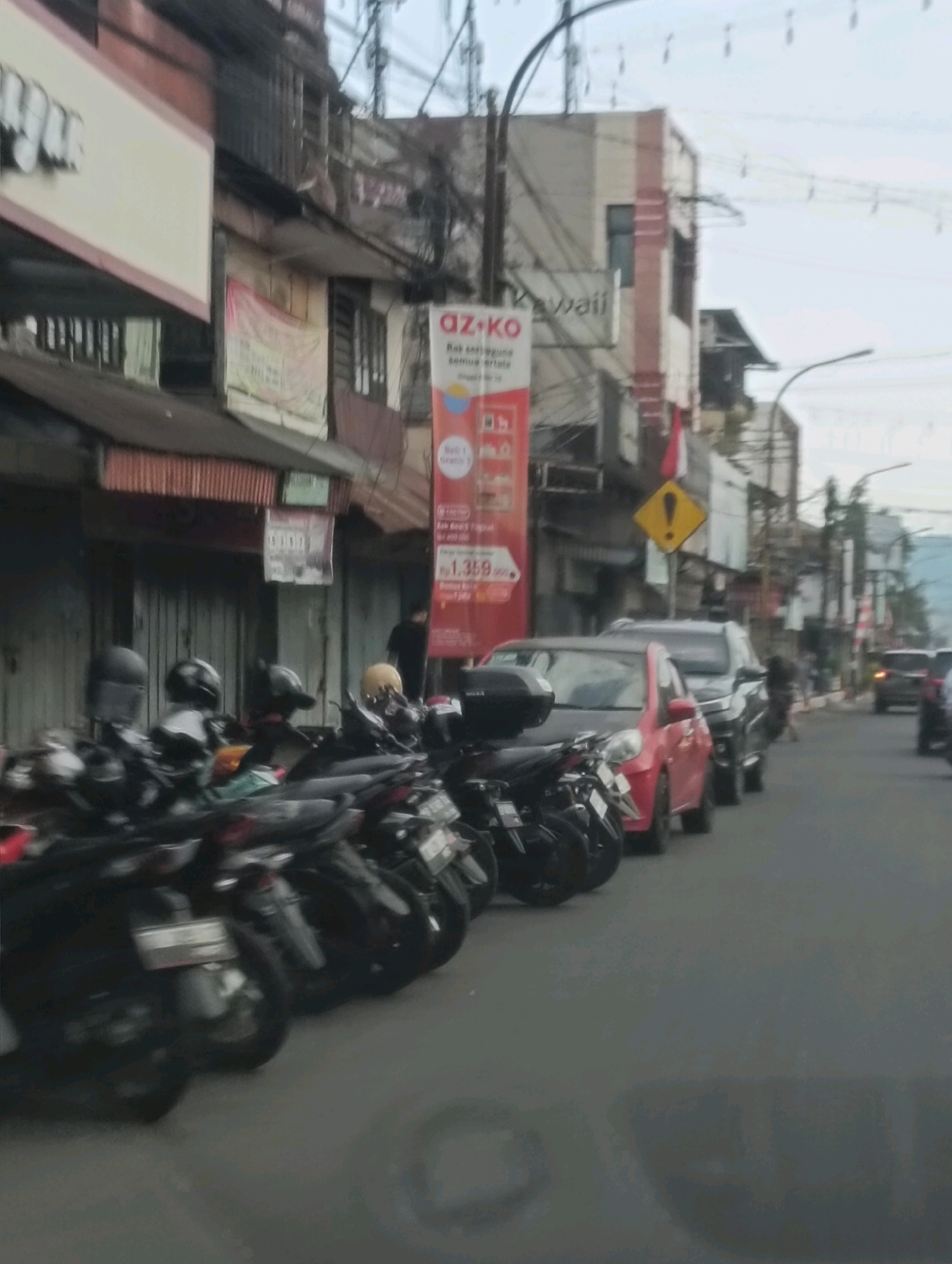 The image depicts a street scene in Tomohon, Indonesia, likely a commercial area. In the foreground, a row of parked motorcycles is visible, with their handlebars and wheels partially obscuring the view. Behind the motorcycles, several cars are parked along the street, including a red car and a dark SUV. The buildings lining the street are multi-story structures with shopfronts and windows. One building has a vertical banner advertising "AZ-KO" with pricing information. The sky is overcast. A yellow diamond-shaped warning sign with a black exclamation mark is mounted on a pole. There are power lines and utility poles visible in the background. The overall impression is of a typical urban street with shops, vehicles, and infrastructure.