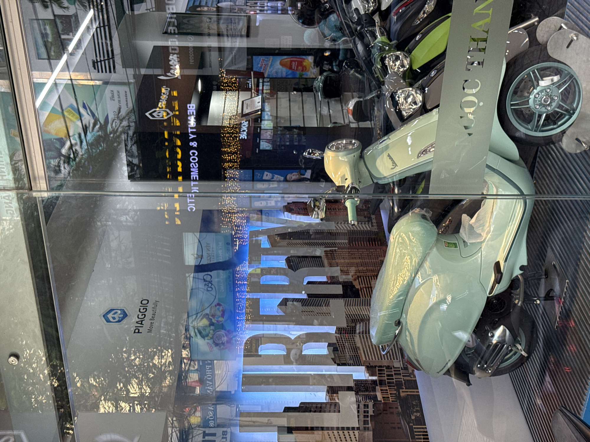 The image shows a storefront display featuring several Piaggio scooters, including a light green model prominently in the foreground. The scooters are positioned behind a glass window, which reflects the surrounding environment, including trees and streetlights. The storefront has large text on the window reading "LIBERTY" in bold, stylized letters against a backdrop of city skyscrapers. Below the "LIBERTY" text, a strip of frosted glass displays text that appears to be in Vietnamese and reads "NGỌC THANH", suggesting the name of the dealership or establishment. Other visible signs and advertisements in the window indicate the sale of beauty and cosmetic products, alongside the scooters. The overall impression is of a modern retail space in an urban setting, likely a showroom for vehicles and related accessories. Based on the visual cues, such as the style of architecture and the presence of Vietnamese text, the location is identified as Nha Trang, Vietnam.