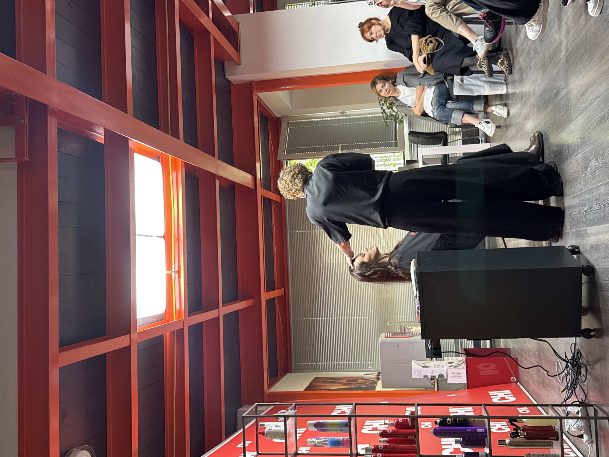 A hair salon scene is captured, with a focus on a hair stylist attending to a seated client. The stylist, wearing a black outfit, is seen working on the client's hair. The setting is in Tbilisi, Georgia, discernible from the specific products visible on a display shelf that prominently features a red CHI product line. In the background, two women are seated in a waiting area; one woman is wearing jeans and a white t-shirt, while the other is in a black top. The location is identifiable as a salon due to the equipment and products displayed, including a black rolling cart positioned near the stylist and the extensive range of hair care products on the shelves. The interior architecture includes a striking, large orange shelving unit.