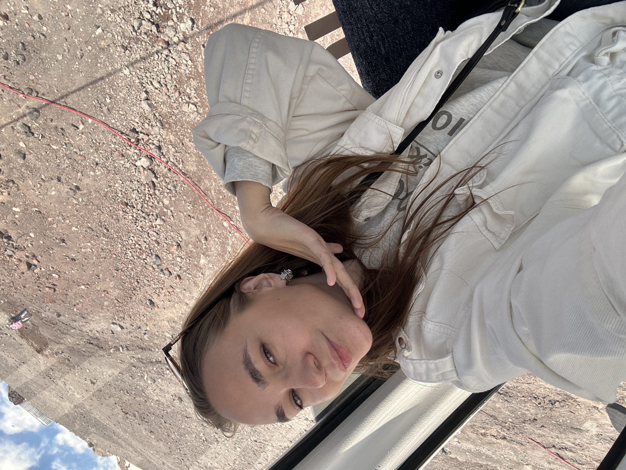 In Terskol, Russia, a young woman with long brown hair is taking a selfie outdoors. She is wearing a white jacket and a grey shirt underneath. Her left hand is resting on her chin, and she is looking at the camera with a slight smile. Sunglasses are perched on top of her head. The background is a steep, rocky hillside with patches of dirt and some sparse vegetation. A red cable runs diagonally across the scene. The sky is visible in the upper left corner, with a few clouds. In the distance, a person in a light-colored outfit and a pink backpack can be seen on the hillside. The overall impression is of a casual outdoor setting, likely in a mountainous or hilly region.