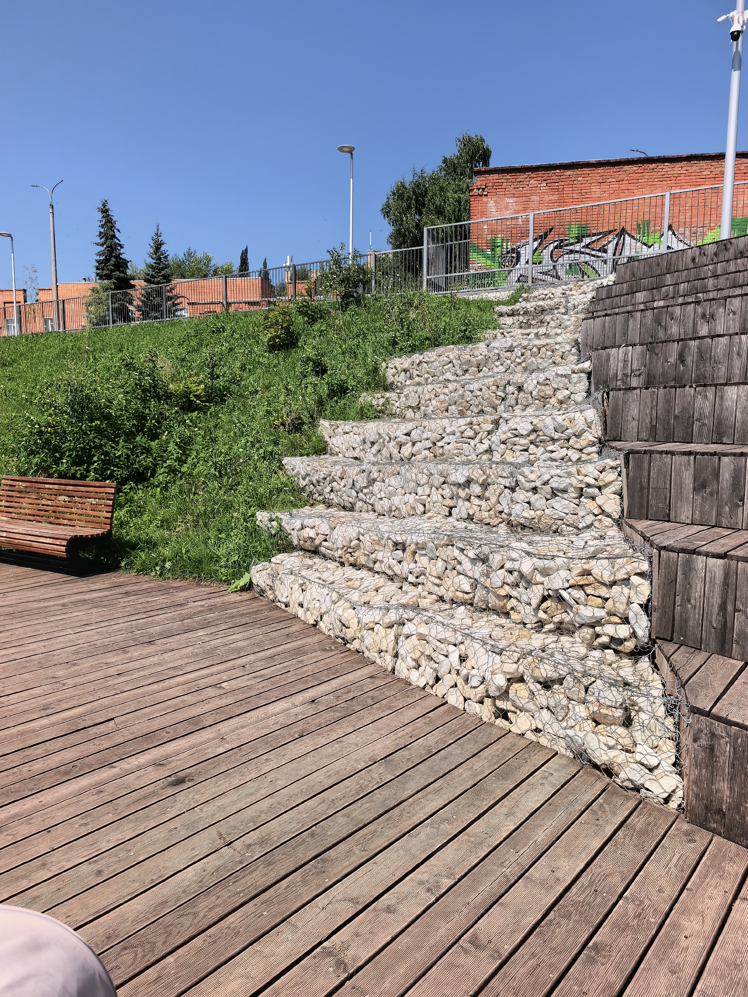 The image depicts a public outdoor space with a wooden deck in the foreground, a gabion staircase filled with stones to the right, and a grassy slope with a wooden bench on the left. In the background, a red brick building with graffiti is visible, bordered by a metal fence and several trees. The sky is clear and blue. This scene is located in Sterlitamak, Russia, likely a park or recreational area. There are no people or animals present in the image.