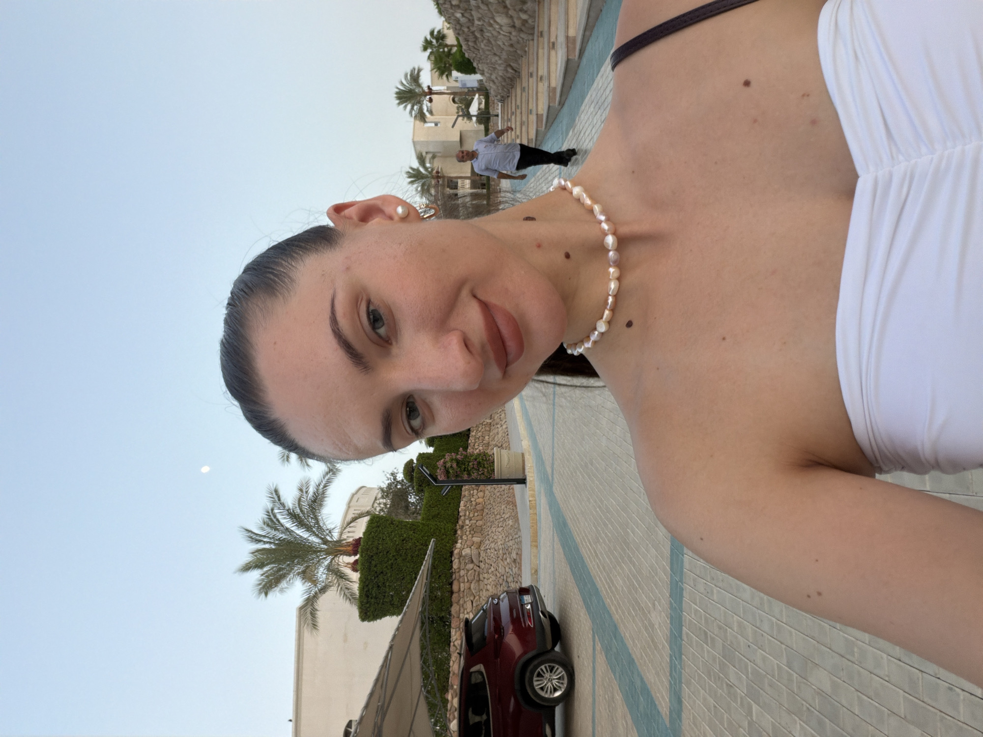 A young woman with her hair pulled back in a ponytail takes a selfie outdoors. She is wearing a white strapless top and a pearl necklace. To her left, a red SUV is parked under a shaded canopy. A man in a light blue shirt and dark pants walks up a set of stairs in the background. Palm trees and buildings are visible in the distance. The scene appears to be in Sharm El Sheikh, Egypt.