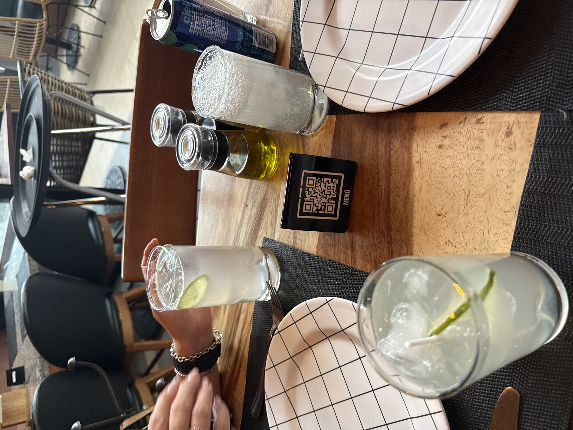 This image depicts a restaurant table in Puebla, Mexico. The table is wooden and features a black placemat underneath two white plates with a black grid pattern. On the table are a can of soda, a glass of salt-rimmed beverage, a salt shaker, an olive oil bottle, and a black card with a QR code that reads "MENU". A glass of a lime-infused drink with ice and a straw is placed on one of the plates, held by a person wearing a black-and-silver bracelet. Another similar drink is visible on the other plate. The surrounding environment shows chairs and other tables, suggesting a restaurant setting.
