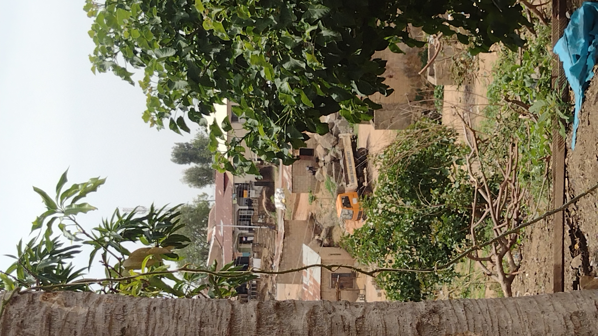 From an elevated vantage point, a scene unfolds within Jos, Nigeria, characterized by modest structures and earthy tones. A weathered wall forms the foreground, providing a textured base from which greenery springs forth—leaves of varying shapes and sizes frame the urban landscape.
Amidst the structures, a construction vehicle, bearing the inscription "KAKI DESIGN INDUSTRY," suggests ongoing development. Piles of what appear to be building materials, including rough-hewn stones, are visible alongside the vehicle. The arrangement of the dwellings and construction suggests a terrain undergoing gradual transformation.
The natural and built environments coexist, with trees and foliage softening the harshness of the construction site. Overhead, a sky shrouded in a thin, diffused light completes the setting.