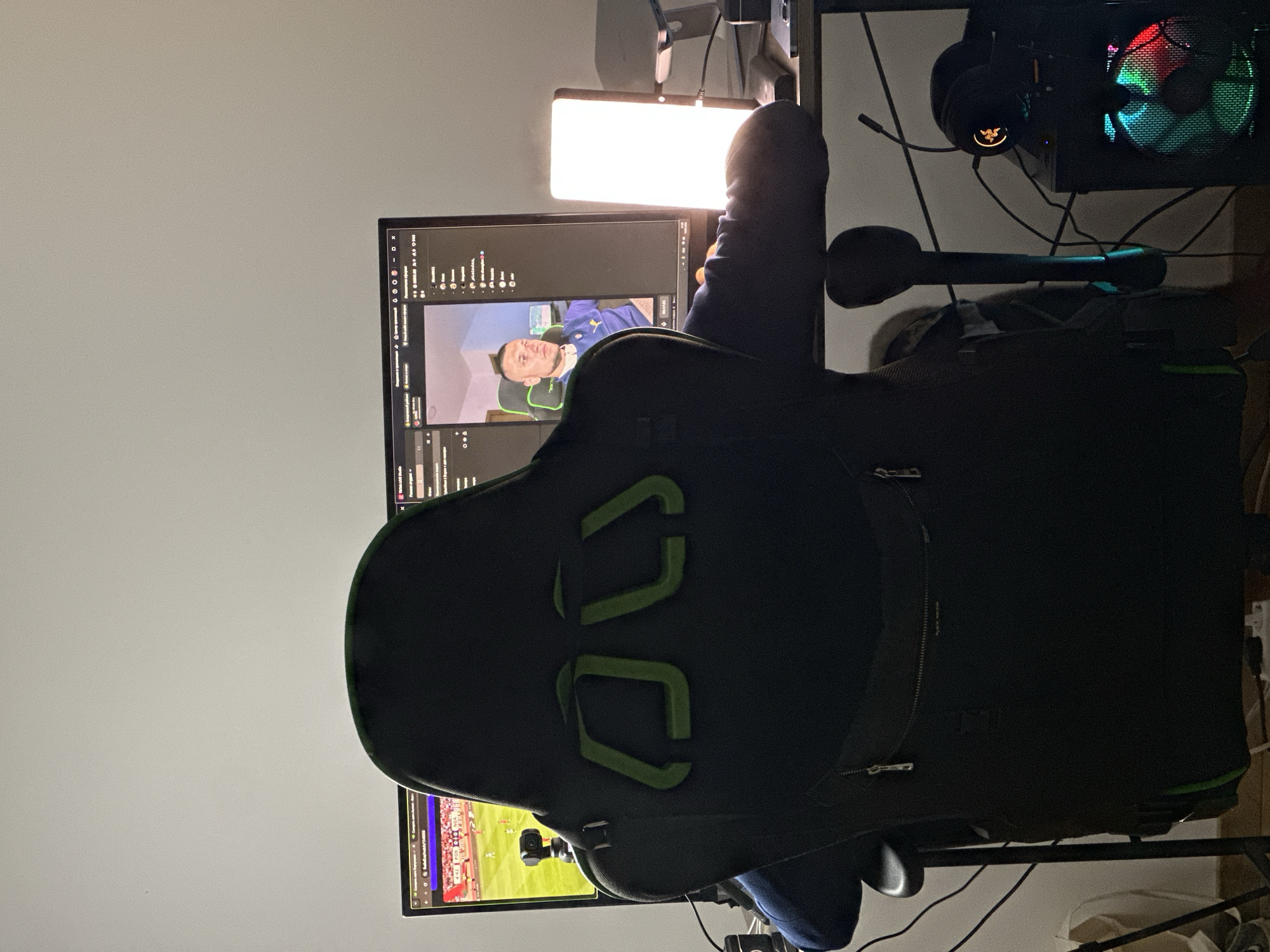 The image depicts a person sitting in a gaming chair, facing away from the camera, and looking at a computer monitor. The monitor displays a split screen: on the left, a sports broadcast showing a soccer match with the score "4:42 POR 0:0 NGA"; on the right, a video stream of a man wearing a blue and yellow shirt. The person in the gaming chair is wearing a dark jacket and blue pants.
In the foreground, the back of the black gaming chair with green accents is prominent. Cables and peripherals are visible around the desk, including headphones with an illuminated logo, a computer tower with a colorful LED fan, and a bright LED light panel. A webcam is positioned on top of the left side of the monitor.
The environment appears to be an indoor setting, likely a room in a home, with a white wall as the backdrop. The lighting is dim, with the primary illumination coming from the monitor and the LED light panel, creating a gaming or streaming setup atmosphere. The location is identified as Donji Pobori, Montenegro.
