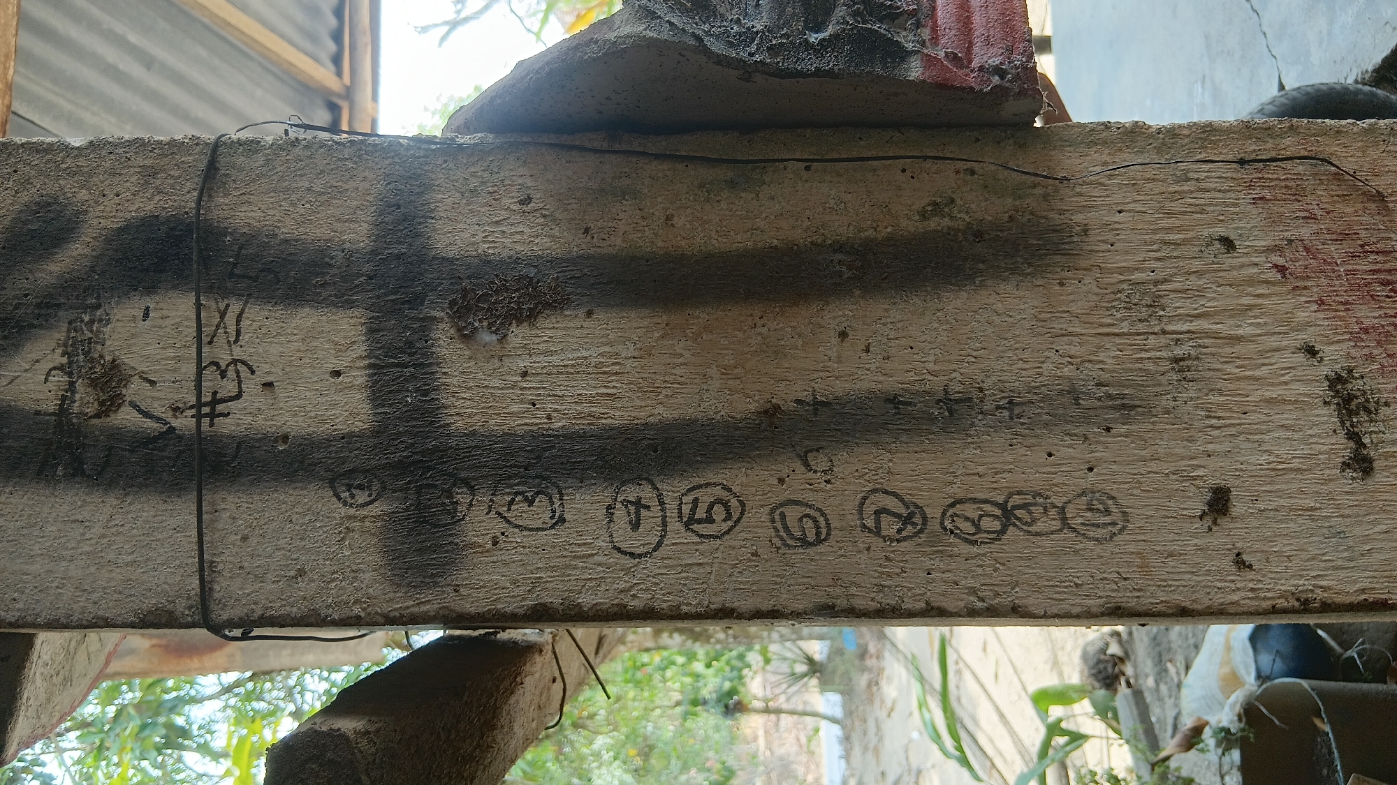 A close-up, eye-level vertical shot shows a weathered, light-brown wooden post in what appears to be an outdoor, possibly rural, setting in Prabumulih, Indonesia. The post is marked with a series of numbers enclosed in circles, ranging from 3 to 10, painted in black ink. Above these numbers, there are some black markings that resemble Asian characters or symbols, alongside a large black spray-painted cross shape. A thin black wire is wrapped around the post. The background is blurred, showing green foliage, a corrugated metal roof, and a hint of a red object on the right side. The overall impression is of a rough, utilitarian structure, possibly part of a fence, gate, or construction site.