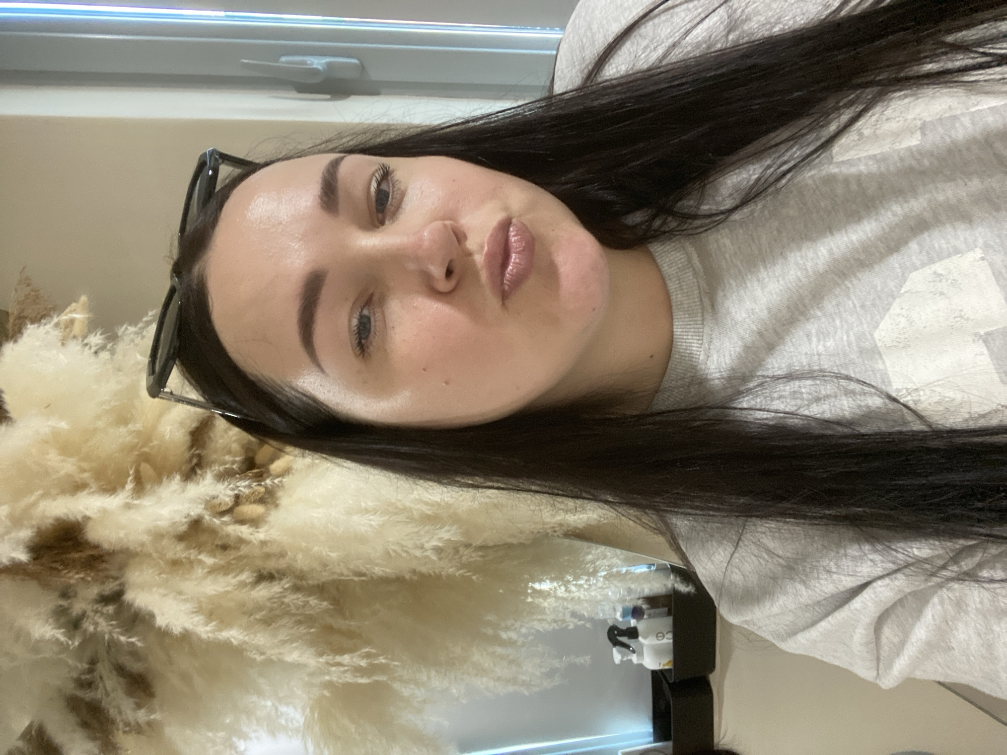 A woman with long, dark hair and fair skin is posing for a selfie. She is wearing a light gray sweatshirt and has sunglasses perched on her head. Her lips are slightly pursed in a kissing expression. In the background, there is a fluffy, dried floral arrangement to the left and a window above her. To the right and reflected in a mirror is some type of counter with hygiene product. The exact location is not discernable and there is no information to establish it is in the city of Sochi, Russia.