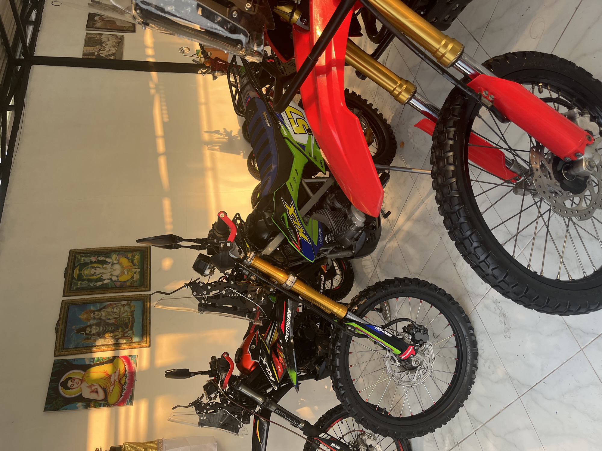 Multiple dirt bikes are parked indoors on a white tiled floor, leaning against a white wall. The scene is illuminated by strong, golden sunlight streaming in from the right, casting long shadows across the floor and highlighting the wall and bikes. The time of day appears to be either morning or late afternoon due to the angle and warmth of the light.
In the foreground, several off-road or dirt motorcycles are visible. The closest bike features a red front fender, gold-colored front suspension forks, and a knobby tire with a disc brake. Another bike, partially visible behind it, has a vibrant green and blue seat and fairings, with text like "Kawasaki" and the number "57" on its side. Further back, another bike with red and black fairings features "FASTHOUSE" branding and "RALLY" on its suspension. Details like "NORIFUMI" and "DID" are also visible on various parts of the bikes, along with tire specifications "80/100-21" on a tire sidewall. The bikes display characteristics typical of dirt bikes, including robust frames, high ground clearance, and specialized tires for off-road use.
On the white wall, three framed religious pictures are hung vertically. From top to bottom, they depict the Hindu deity Ganesha, followed by Shiva and Parvati, and finally, Buddha in a meditative pose. The sunlight directly illuminates these pictures, enhancing their colors and details. The metal framework of what appears to be a roof support or overhead structure is visible on the far left. The overall setting suggests a personal garage or a display area for these motorcycles, possibly in Tegallalang, Indonesia, given the cultural context of the artwork and the general tropical lighting.