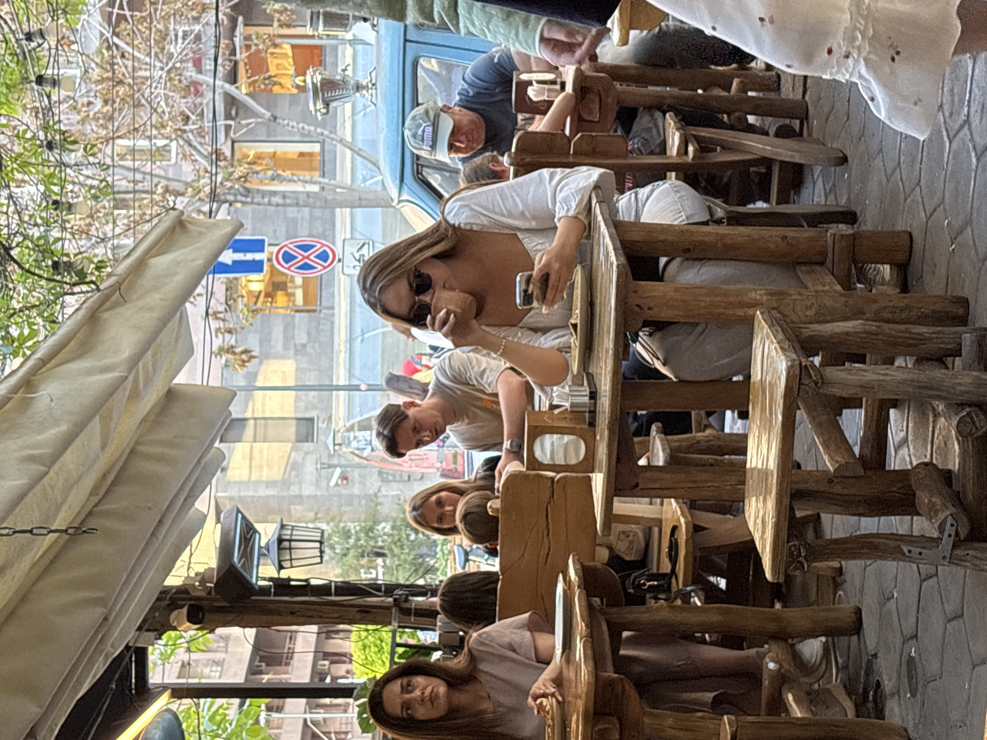 The image shows a street cafe scene in Yerevan, Armenia, with several people seated at rough-hewn wooden tables and chairs under a canvas awning. A woman in a white top and sunglasses is holding a mobile phone to her ear, while another person in a grey shirt sits at the next table. Further along the row, other individuals are visible, with a man in a Puma cap seated in the background near a vintage car. In the background, there is a building with street signs, including a no-parking sign. The scene conveys a casual, outdoor dining atmosphere.