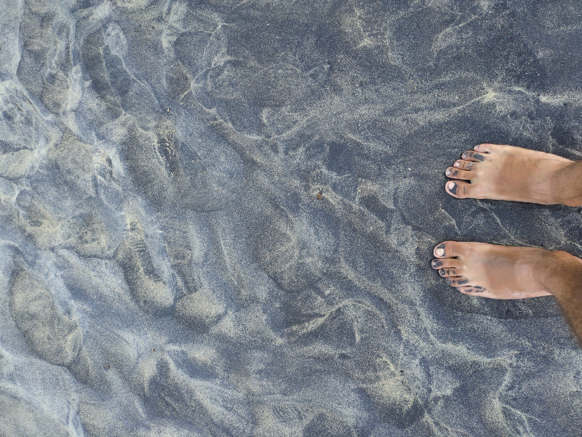 An overhead photograph captures a pair of human feet standing on a beach in East Quy Nhon Ward, Vietnam. The sandy surface beneath the feet displays a natural blend of dark gray-black and lighter beige sand, intermixing to create variegated patterns. Both feet are bare, with dark sand particles visible adhering to the skin, primarily around the toes and underneath the toenails. The skin exhibits a light to medium brown tone, and some hair is present on the lower legs, extending slightly into the frame from the bottom edge.