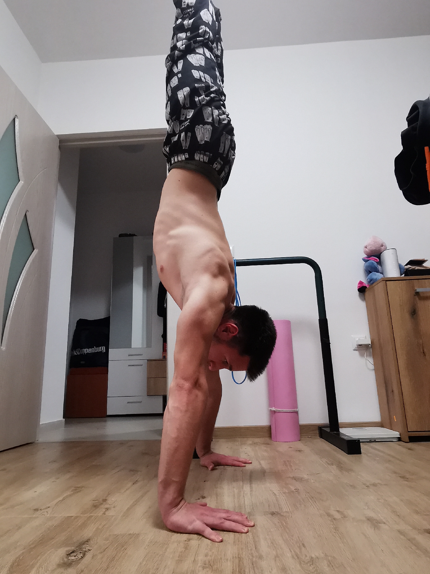 A man is performing a handstand indoors. He is wearing black and white patterned pants and appears to be shirtless. His hands are flat on a wooden floor. A doorway is visible to his left, leading to another room with furniture. To his right is exercise equipment, a rolled-up pink yoga mat, a scale, and a wooden cabinet with items on top, including a stuffed animal and a white cylindrical object. A black hoodie is hanging from the cabinet. The walls are white. The location is an interior setting. There is no indication of Burgas, Bulgaria.