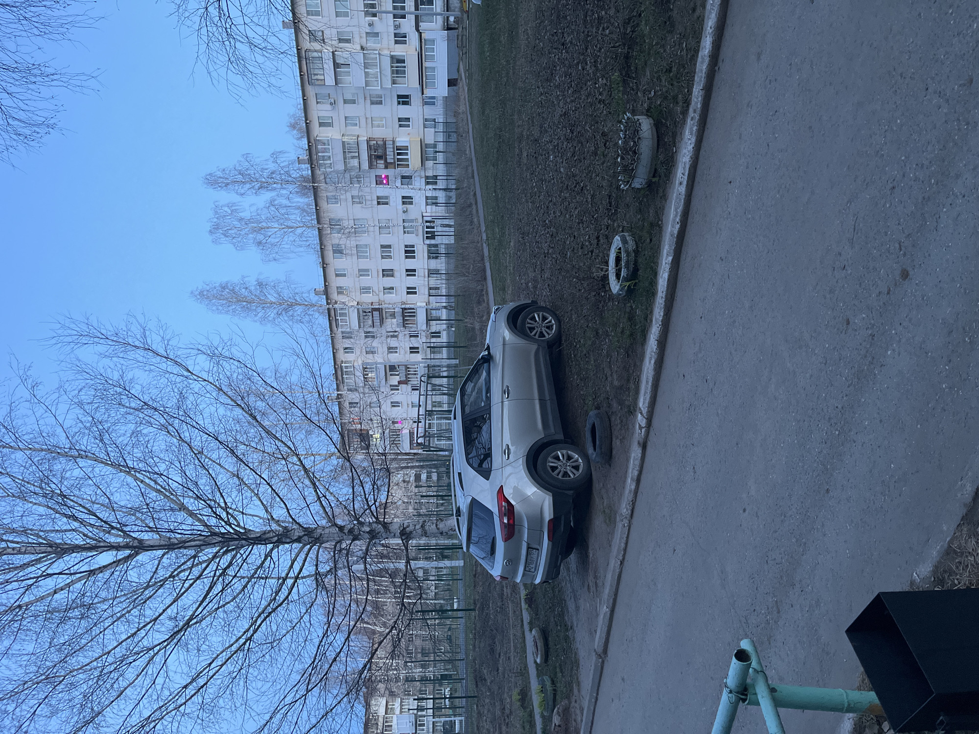 The image presents an outdoor residential scene, likely during late winter or early spring, under a bright, clear blue sky. No people or specific activities are visible, conveying a sense of quiet daily life.
In the background, a large, multi-story apartment building with numerous windows and balconies stretches horizontally across the frame. Its architecture is typical of residential buildings found in post-Soviet urban areas. A distinct pink light emanates from a window on an upper floor of the building. Several bare deciduous trees, their branches devoid of leaves, stand between the viewer and the apartment block, with more leafless trees visible further down the street.
The foreground features a paved asphalt path or sidewalk along the right side, separating it from a grassy area to the left. Parked partially on this grassy strip and close to the path is a silver or light-colored SUV, specifically a Hyundai Creta. Its rear license plate, "H248BY 159", is visible, indicating Russian registration from the Perm Krai region.
Also on the grassy area, two old car tires are repurposed, seemingly as decorative borders or planters; one is painted white and partially filled with dry vegetation, while the other is unpainted. In the bottom right corner, a black square-shaped trash bin or container is partially visible, along with part of a light blue metal structure, possibly belonging to playground equipment. The scene captures an ordinary urban landscape.
