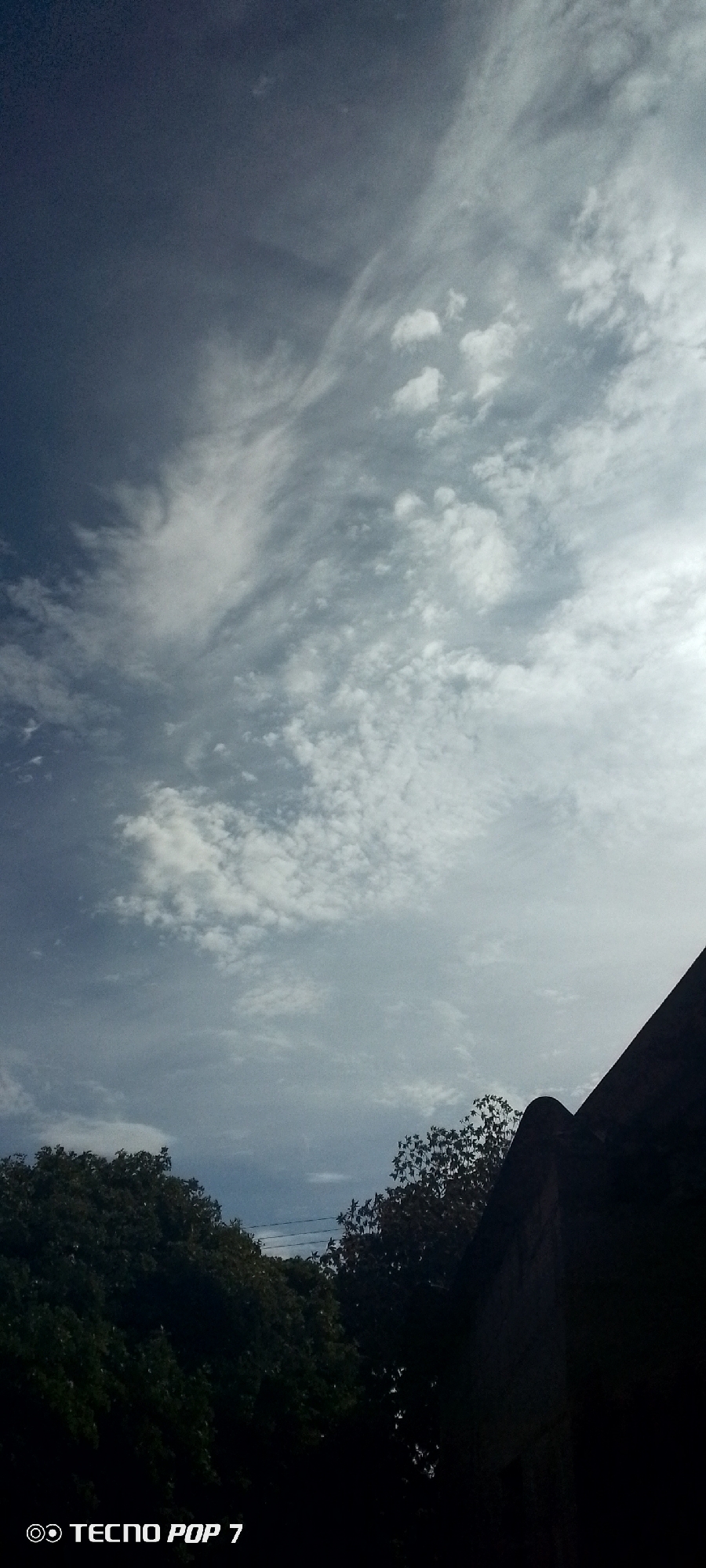 The image captures a view of the sky in what appears to be the city of Altagracia, Venezuela, characterized by altocumulus clouds against a backdrop of varying shades of blue. Below the sky, vegetation, potentially trees, is visible on the lower left and center. On the lower right, there is a dark, undefined structure that could be part of a building. The image includes a watermark on the bottom left corner indicating it was taken with a TECNO POP 7.
