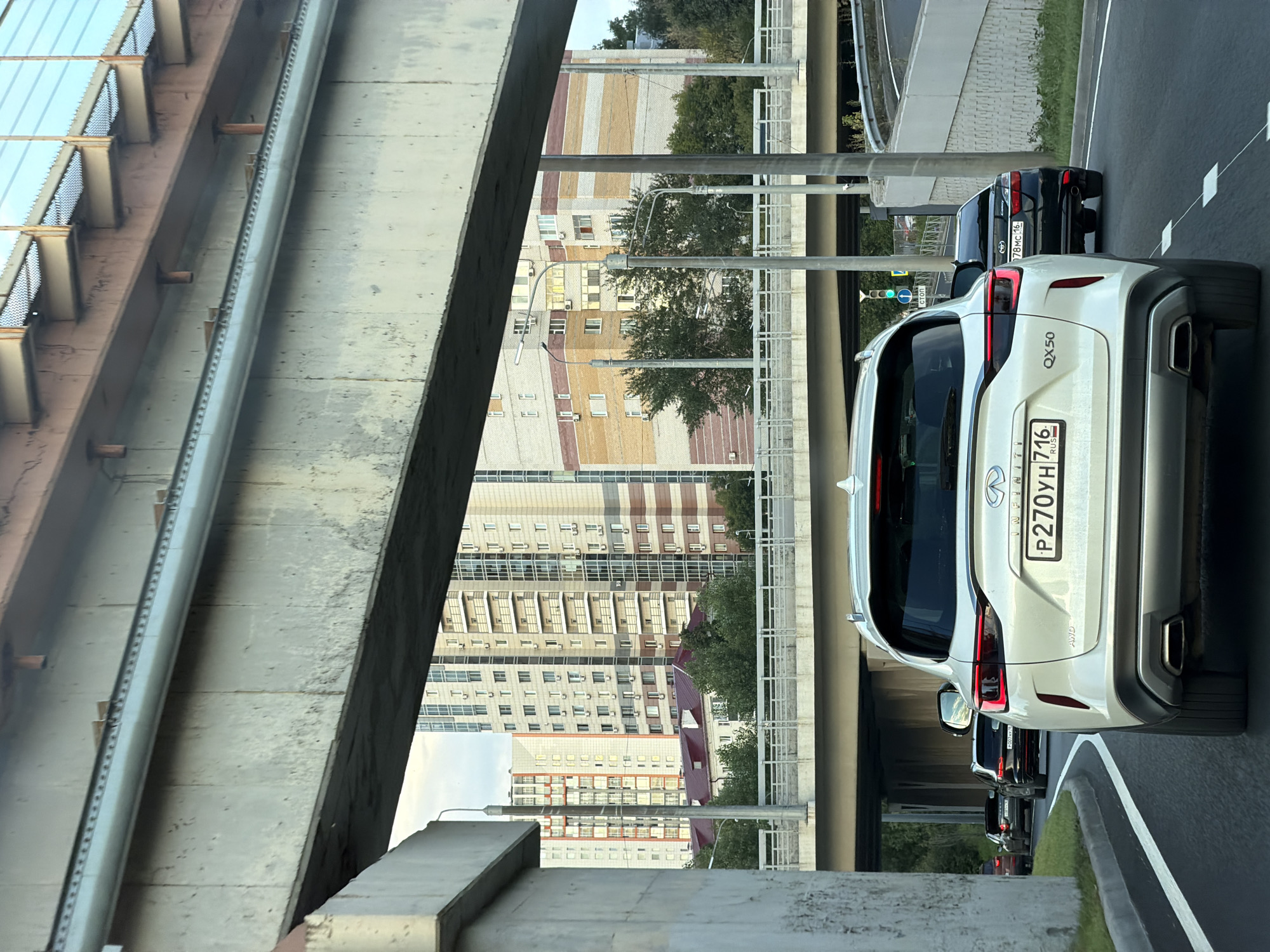 An eye-level, medium shot captures a white Infiniti QX50 SUV in the foreground, its rear facing the camera and angled slightly to the right. The car's license plate reads "P270YH 716 RUS". To its right, a black sedan is visible, and further back, other cars are seen in traffic on a multi-lane road. The road is flanked by concrete barriers and white lane markings. Overhead, a large concrete overpass or bridge dominates the upper portion of the frame, with structural details and a metallic element visible. Beyond the overpass, multi-story residential buildings with a mix of brick and painted facades are clustered together, some featuring balconies and numerous windows. Trees and streetlights are also present in the scene. The overall setting appears to be an urban environment with elevated roadways and high-rise architecture.