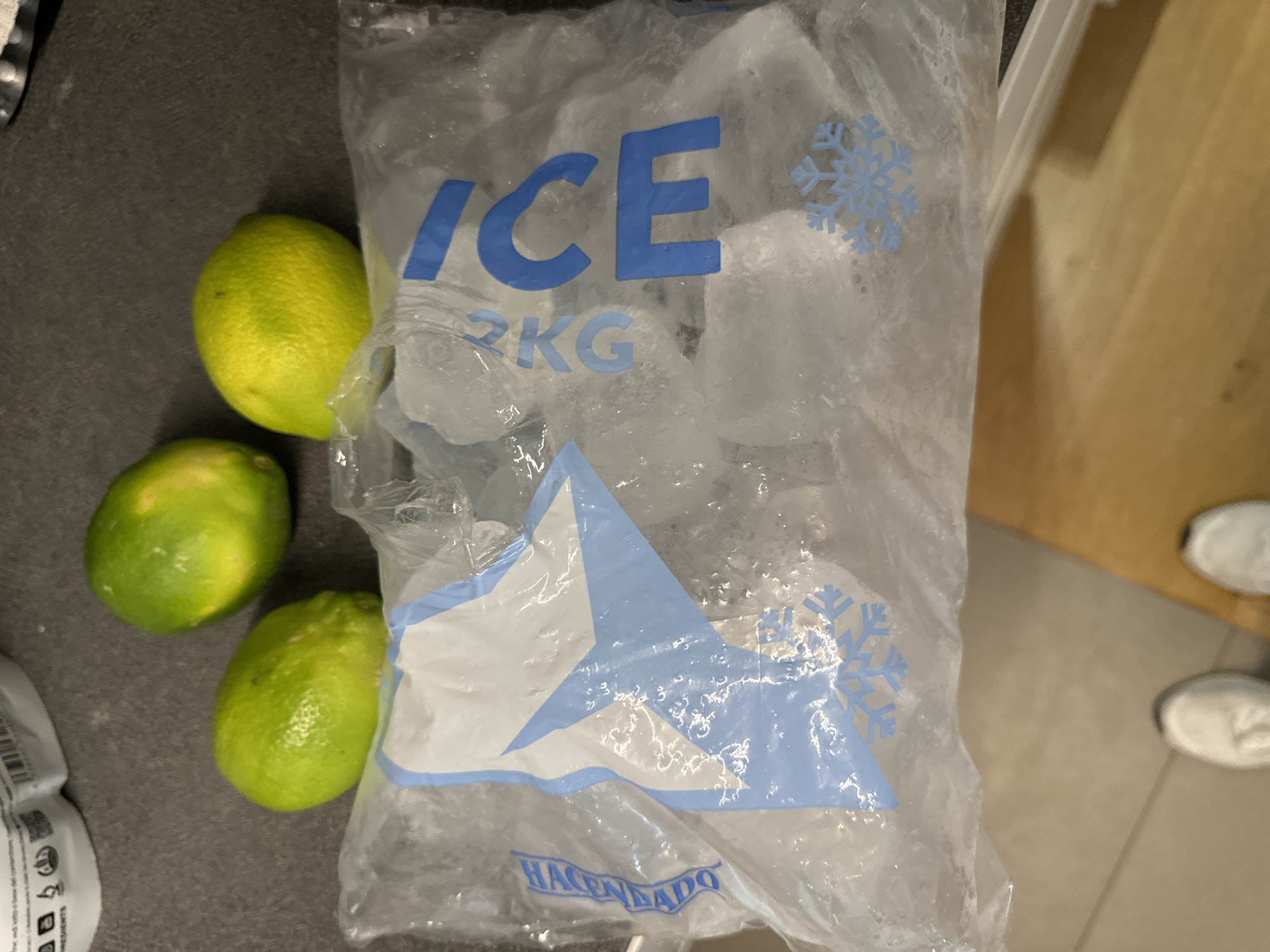 The image shows a bag of ice cubes next to three limes on a dark gray surface, likely a countertop. The bag of ice has blue text that reads "ICE" and "2 KG". There is also a blue snowflake design on the bag. A blue and white star design is visible on the lower part of the bag, with the word "HACENDADO" in blue text below it. The limes are green and appear to be fresh. In the background, a wooden surface is visible to the right, and white shoes can be seen on a tiled floor in the far background. The lighting suggests it is daytime. There are no people visible in the foreground, but the presence of shoes in the background might imply someone is nearby. The scene appears to be an indoor kitchen or food preparation area.