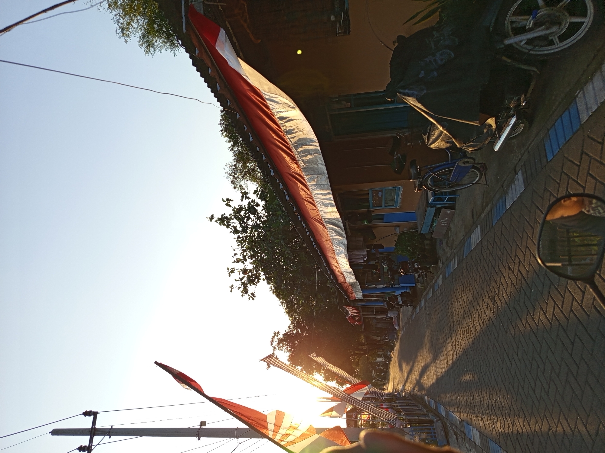 The image depicts a street scene in Tebel, Indonesia, during what appears to be sunrise or sunset, with the sun low on the horizon casting long shadows. The street is paved with interlocking brick pavers, bordered by a blue and white stripe. On the left side of the street, a utility pole stands tall, with electrical wires stretching across the frame. Several Indonesian flags, red and white, are displayed on poles along this side, some adorned with checkered patterns, and one is illuminated by the sun.
To the right of the street, a row of single-story houses lines the path. These houses have corrugated metal roofs and are painted in warm, earthy tones. Red and white awnings are stretched over the front of some of the houses, also reminiscent of the Indonesian flag. Various objects are visible in front of the houses, including bicycles and motorcycles. One motorcycle is covered with a black tarp. A blue wooden bench is placed in front of one of the doorways.
The environment appears to be a residential area within a city or town, with vegetation visible behind the houses. The lighting suggests a quiet time of day, possibly early morning or late afternoon, with a clear sky. No people are clearly visible or engaged in any specific actions. The image is taken from a low vantage point, possibly from a vehicle or looking over handlebars, as suggested by the reflection in the lower right corner.