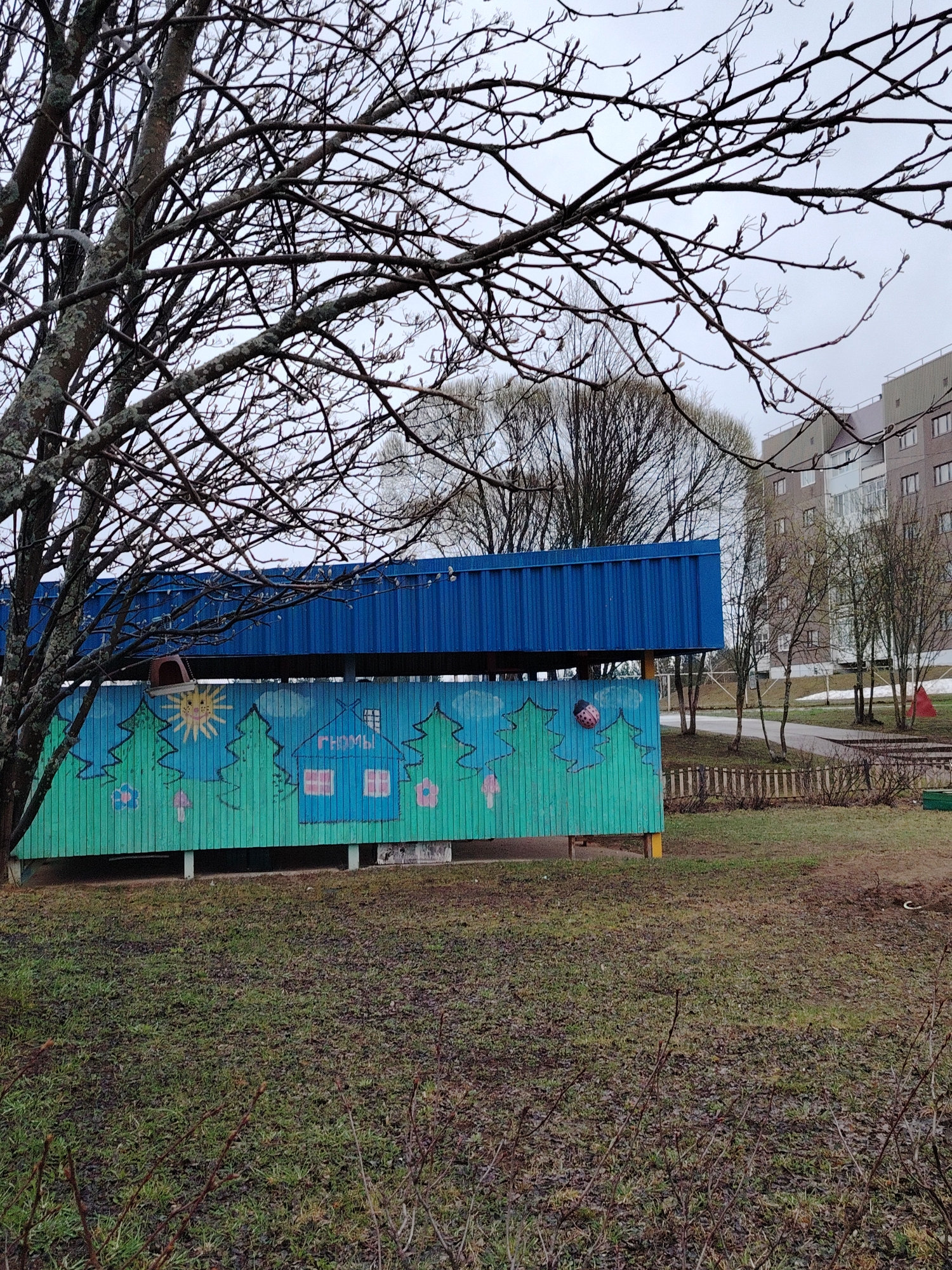 The image captures an outdoor area, likely a playground or kindergarten yard in Марковский, Russia, on an overcast day, suggesting early spring or late autumn. A large deciduous tree with bare, lichen-covered branches and emerging buds fills the left foreground, its canopy extending across the top of the frame. A small, dark bird feeder hangs from one of its branches.
Central to the scene is a two-part outdoor structure. The upper section is a bright blue corrugated metal roof or wall, while the lower section features a vibrant mural painted on vertical wooden planks. The mural illustrates a cheerful landscape with stylized green evergreen trees, a yellow sun with a smiling face, pink and blue flowers, and mushrooms. A blue house with red-framed windows is painted prominently, bearing the white Cyrillic text "ГНОМЫ" (Gnomes) on its facade. A three-dimensional red and black ladybug decoration is attached to the right side of the painted trees.
Behind this painted structure, a low wooden fence is visible, separating it from a larger grassy expanse. Several bare trees with slender trunks are scattered in the mid-ground, and in the background, a multi-story residential building with multiple windows and balconies rises under the grey sky. The ground throughout the image is a mix of sparse, wet grass and darker patches of earth, consistent with temperate weather before full spring bloom. Some red geometric shapes, possibly playground elements, are faintly visible in the far right background. No direct human activity is observed.