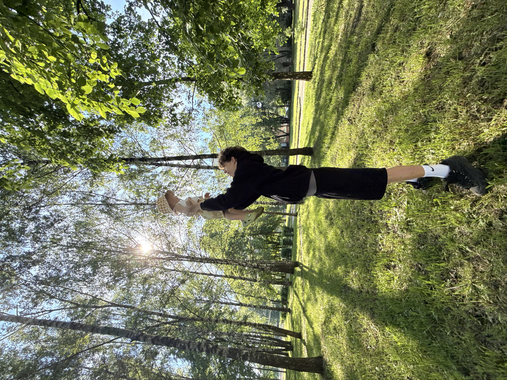 In a sun-dappled park with tall birch trees and lush green grass, a young person in a black hoodie and shorts is holding a baby aloft. The baby is wearing a checkered hat and light-colored clothing. Sunlight streams through the leaves of the trees, casting dappled shadows on the ground. The background shows more trees and a hint of residential buildings and a road. The overall scene depicts a peaceful moment of play between an older sibling or parent and a baby in a natural outdoor setting.