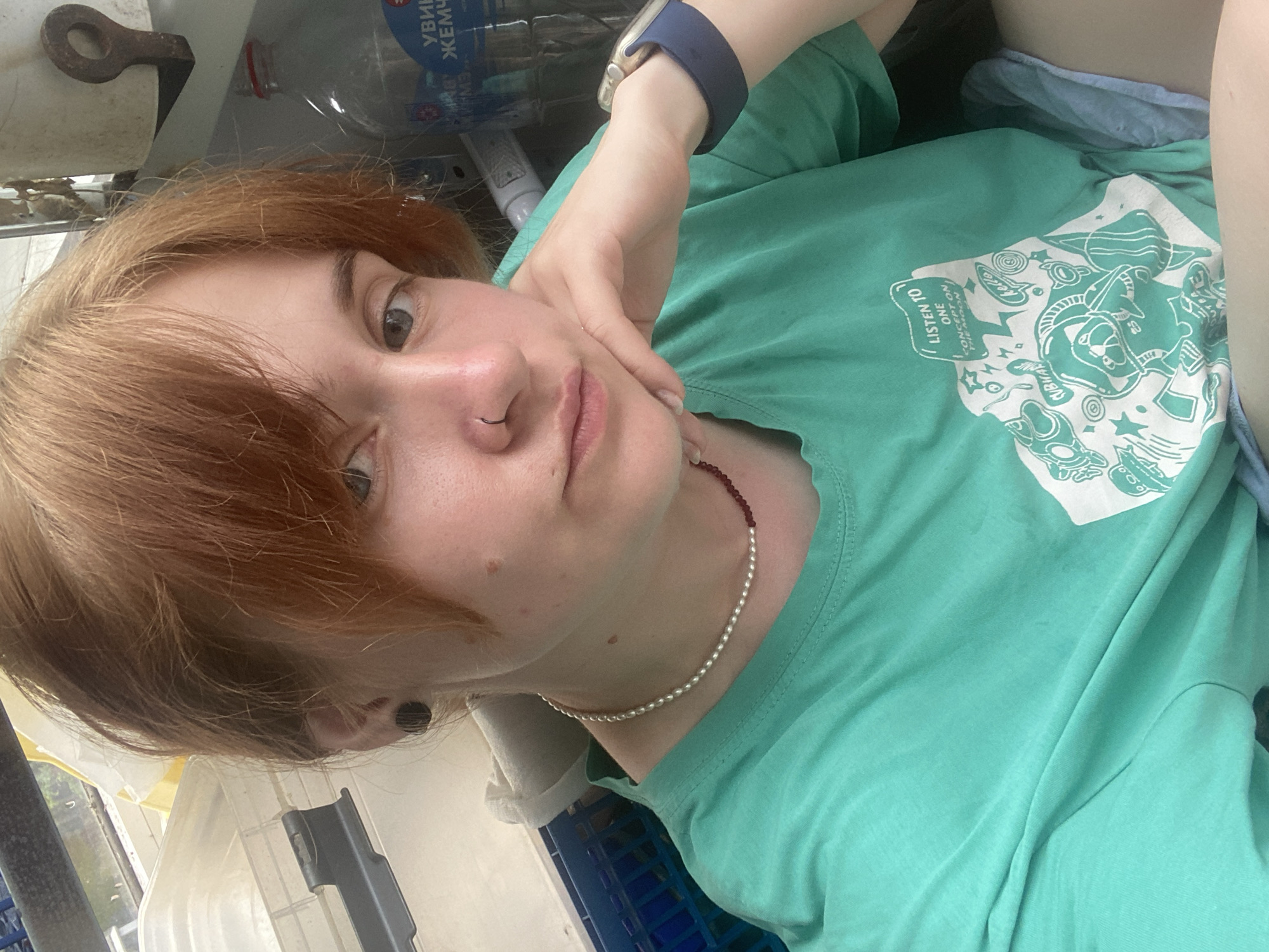 A young woman with reddish-brown hair, wearing a teal t-shirt and blue shorts, poses for a selfie. She has a nose ring and a pearl necklace. Her right hand rests on her chin, and she is wearing a smartwatch on her left wrist. The background appears to be an indoor space with various containers, a bottle of water with Russian labeling, and part of a window. The location is identified as Chaikovsky, Russia.