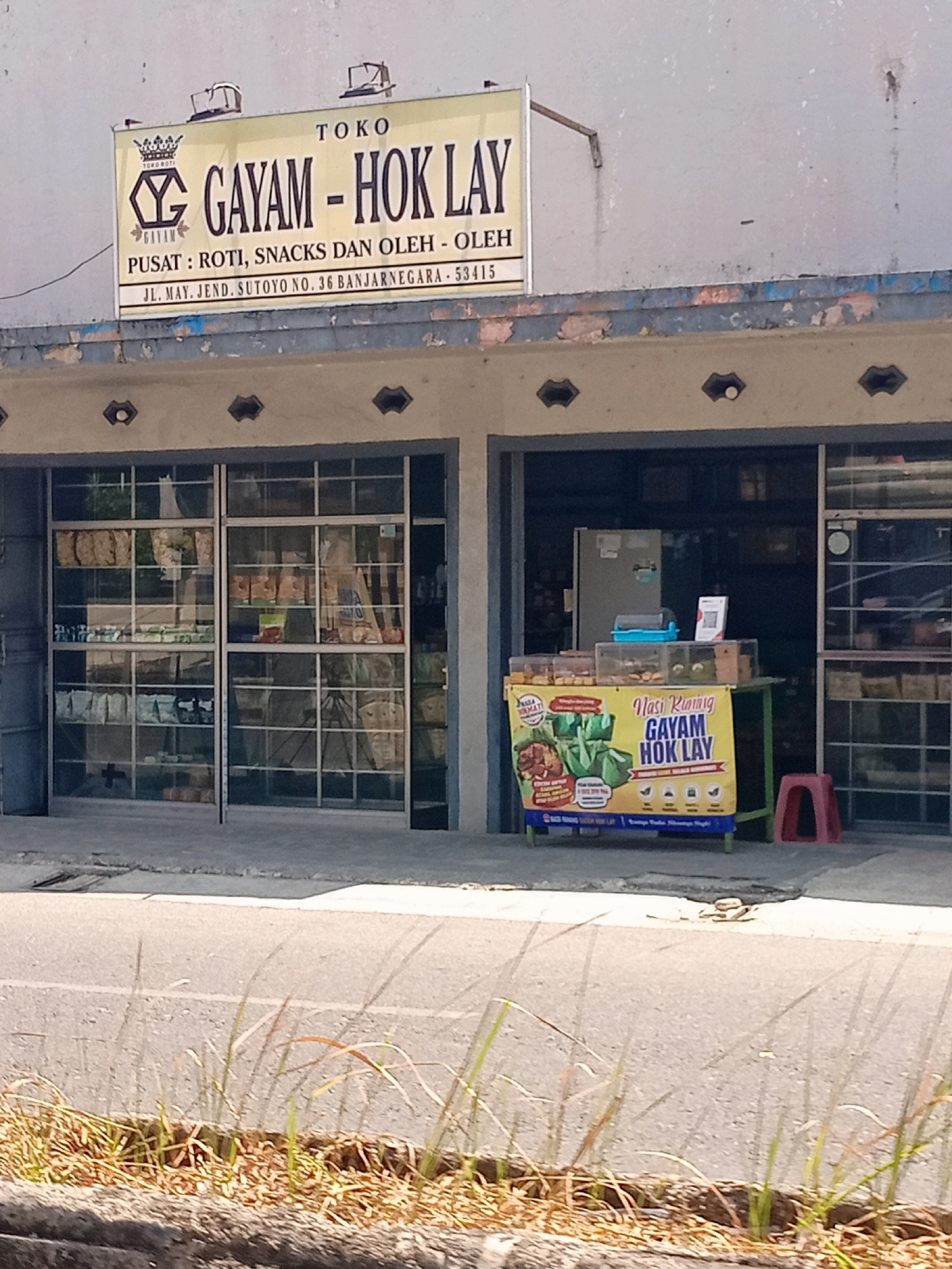 The image shows the storefront of a business named "GAYAM - HOK LAY" located at JL. MAY. JEND. SUTOYO NO. 36 BANJARNEGARA - 53415, Indonesia. The business appears to be a small shop selling roti (bread) and snacks. In front of the shop, there is a yellow banner advertising "Nasi Kuning GAYAM HOK LAY". The storefront has large glass windows displaying various products. A red stool is placed outside the entrance. The street in front of the shop is paved with asphalt and a small patch of grass and dry vegetation is visible in the foreground.
