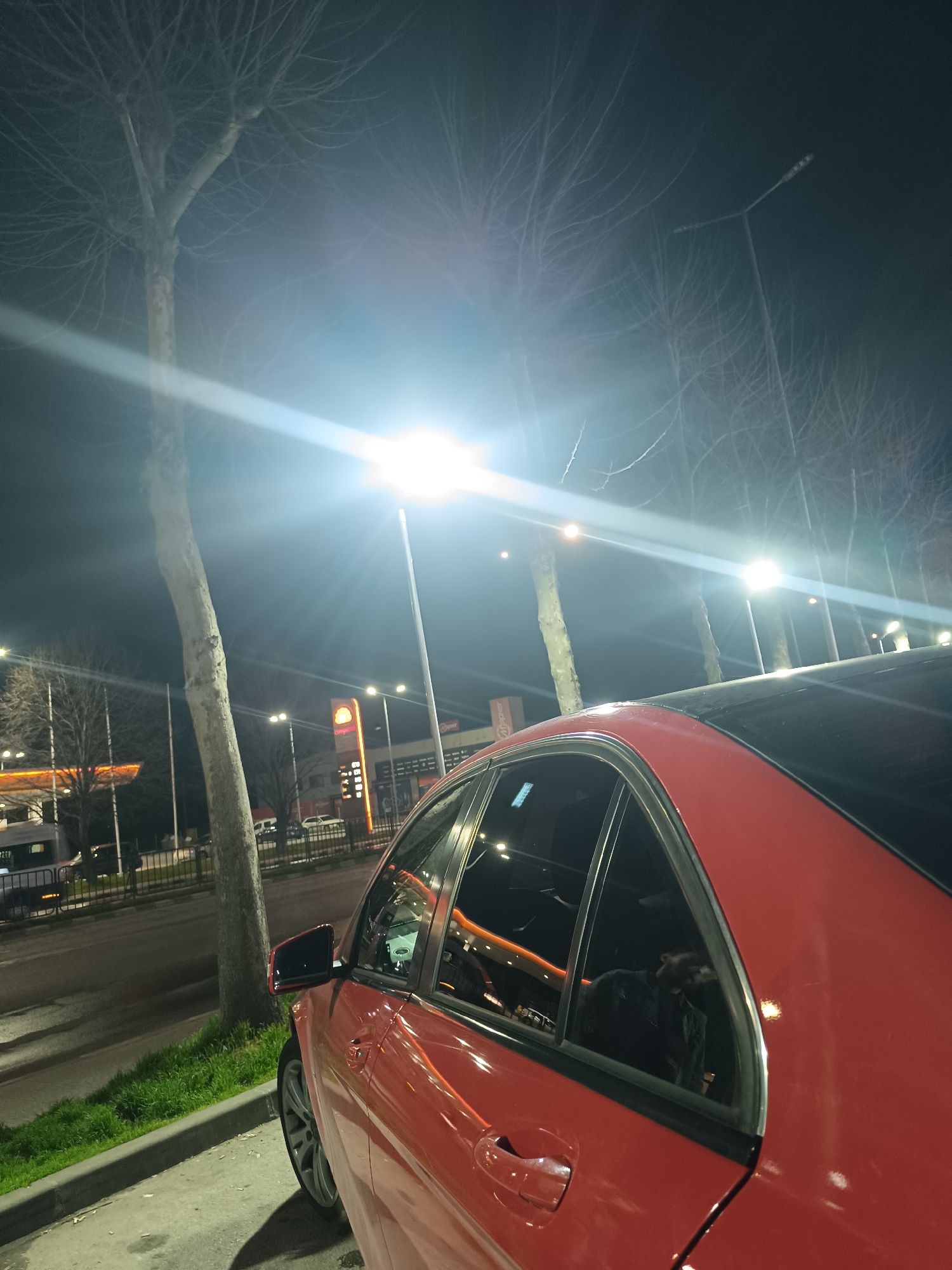 A red car is parked at a petrol station in Asenovgrad, Bulgaria at night. Streetlights illuminate the area and there are bare trees along the road. The reflection of the petrol station and lights are seen in the car windows. A curb and grass are next to the car.