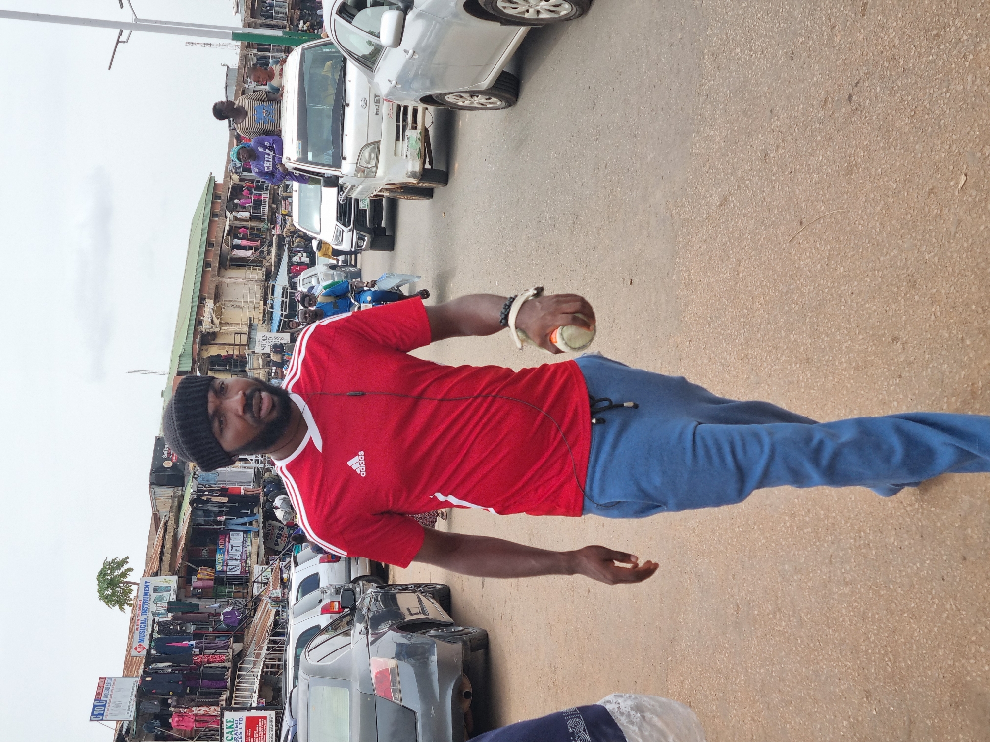 The image depicts a man standing on a street in what appears to be a market area, possibly in Jos, Nigeria, based on the signage and the general environment. The man is wearing a black beanie, a red Adidas t-shirt, and blue jeans. He is holding a tennis ball in his right hand and has what appears to be a beaded bracelet on his left wrist. He appears to be looking directly at the camera.
Behind the man, there is a busy street with several vehicles, including cars and a white HiJET van. Buildings line the left side of the street, housing various shops with signs visible, including "MUSICAL INSTRUMENT," "OLIVE NINE," "CTOC WHOLESALE & RETAIL," and "CAKE GRATED." Clothes are displayed outside of a few of the shops. The overall environment suggests a bustling commercial area.