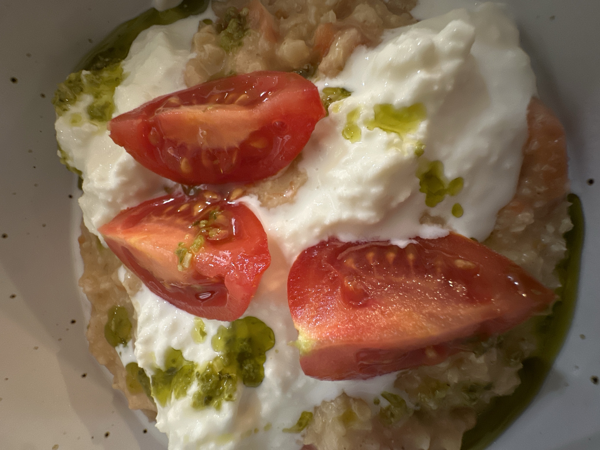 A close-up, overhead shot depicts a food dish, likely served in Novi Sad, Serbia. The dish contains a base of what appears to be coarse bread or couscous, topped with a generous dollop of white cheese (possibly burrata or a similar soft cheese). Three wedges of ripe, red tomato are placed on top of the cheese. Green droplets, potentially pesto or flavored oil, are drizzled across the cheese and the bread base. The food is presented in a shallow, off-white bowl with a hint of speckled texture. No people are visible.