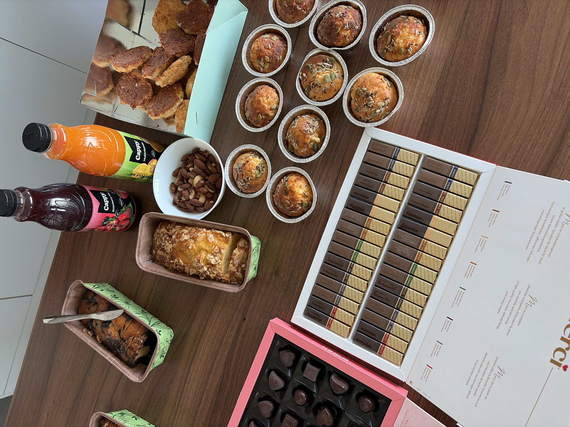 A wooden table displays an assortment of food items. In the foreground left, a pink box contains an arrangement of chocolates, with some candies missing. Next to it, a white box is filled with individually wrapped chocolate bars, many of which are a dark color with gold or multi-colored wrappers. In the upper left corner, a green paper loaf pan holds a partially eaten banana bread with a knife stuck into it. To its right, two glass bottles with black caps are present: one contains a dark red liquid, labeled "Cappy Strawberry Mix," and the other contains orange juice, labeled "Cappy Multivitamin." Between the loaf pan and the bottles, a white bowl holds a pile of almonds. To the right of the almonds, a light blue tray contains several small, golden-brown baked goods topped with seeds, likely muffins or scones. Behind these, another tray holds more of these seed-topped baked goods. Further to the right and slightly out of focus, a light blue box is filled with more of the golden-brown baked goods. The lighting suggests an indoor environment, possibly a kitchen or dining area, with the wooden table surface being the dominant element of the environment.