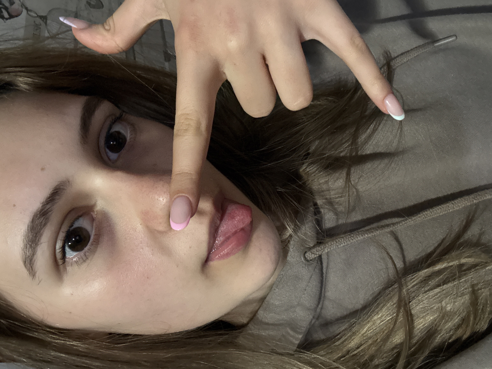 The image captures a close-up, rotated portrait of a young person, likely female, from a slightly elevated perspective. The person has long, light brown hair that frames their face and spreads around them. They are wearing a grey or earthy-toned hooded sweatshirt, with the drawstrings of the hood visible near their right shoulder.
Their face is positioned towards the center-left of the frame, with their dark brown eyes looking directly at the viewer. Their skin appears clear with a few small moles or freckles visible. The person's tongue is slightly extended from their mouth in a playful manner. A hand is prominently featured across their face, with fingers spread out. The index finger points towards the right side of the frame, and the pinky finger lightly touches the bridge of their nose.
A notable detail is the manicure on the visible nails. The nail on the index finger features a light blue or mint-colored French tip, while the pinky finger's nail has a light pink French tip. Another nail, partially visible further up on the hand, appears to have a classic white French tip.
The background is indistinct but suggests an indoor setting, possibly a bed or sofa, with faint patterns visible in the top left corner of the original image. The lighting is soft and ambient. The overall impression is casual and playful, with the pose and expression conveying a sense of lightheartedness. The image is significantly rotated, making the person appear to be lying on their side or back.