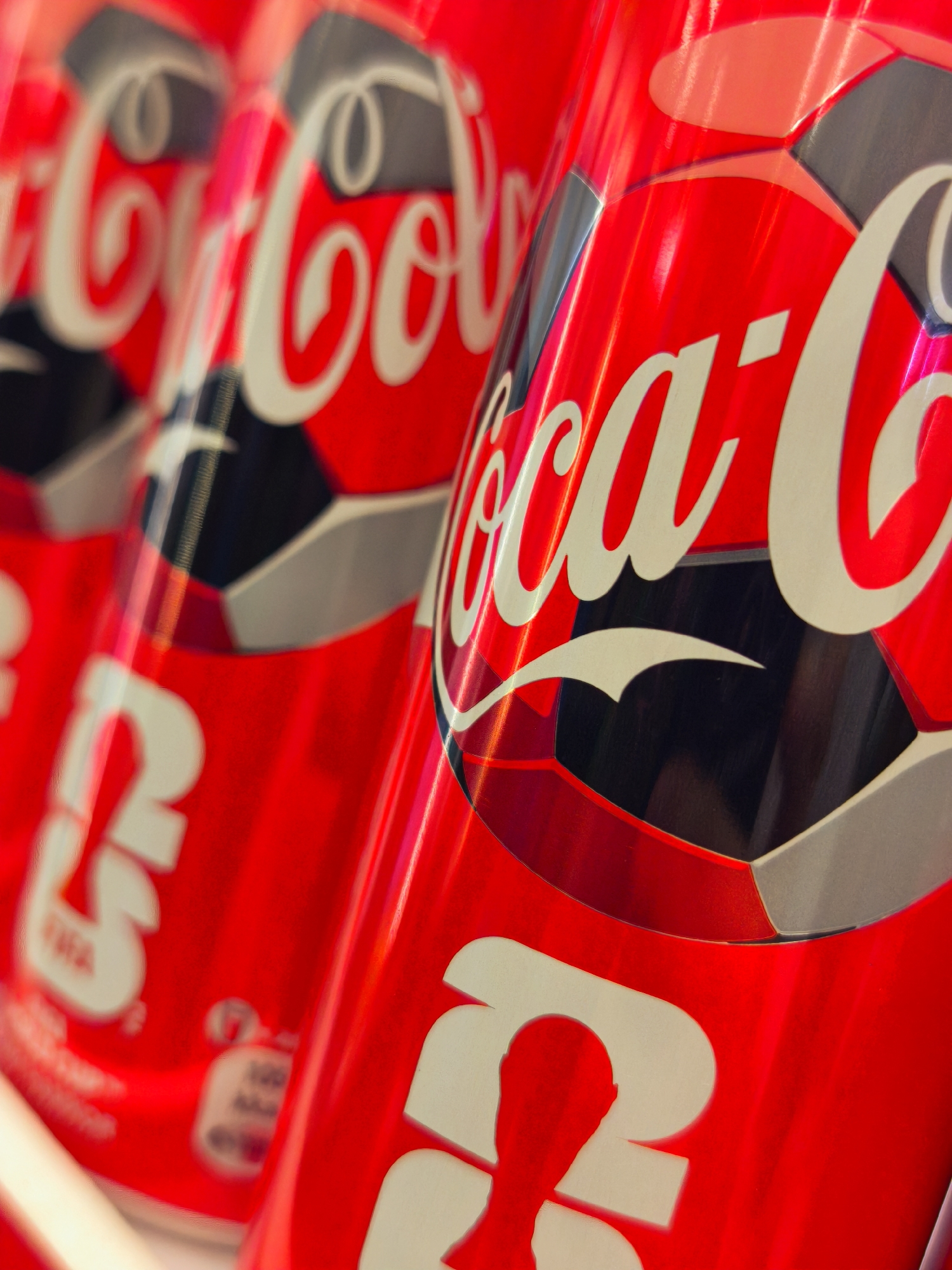 The image displays a close-up, angled view of several red Coca-Cola cans, some with white lettering that reads "Coca-Cola" in a script font. The cans appear to be stacked or arranged in a line. The cans have a metallic sheen and are reflecting light. There are also soccer ball-like patterns in black and gray on some of the cans. The background is out of focus and appears to be a commercial setting, possibly a store or a display. The image does not provide any information or visual cues that would allow for the determination of its location in Tashkent, Uzbekistan, or any specific city.
